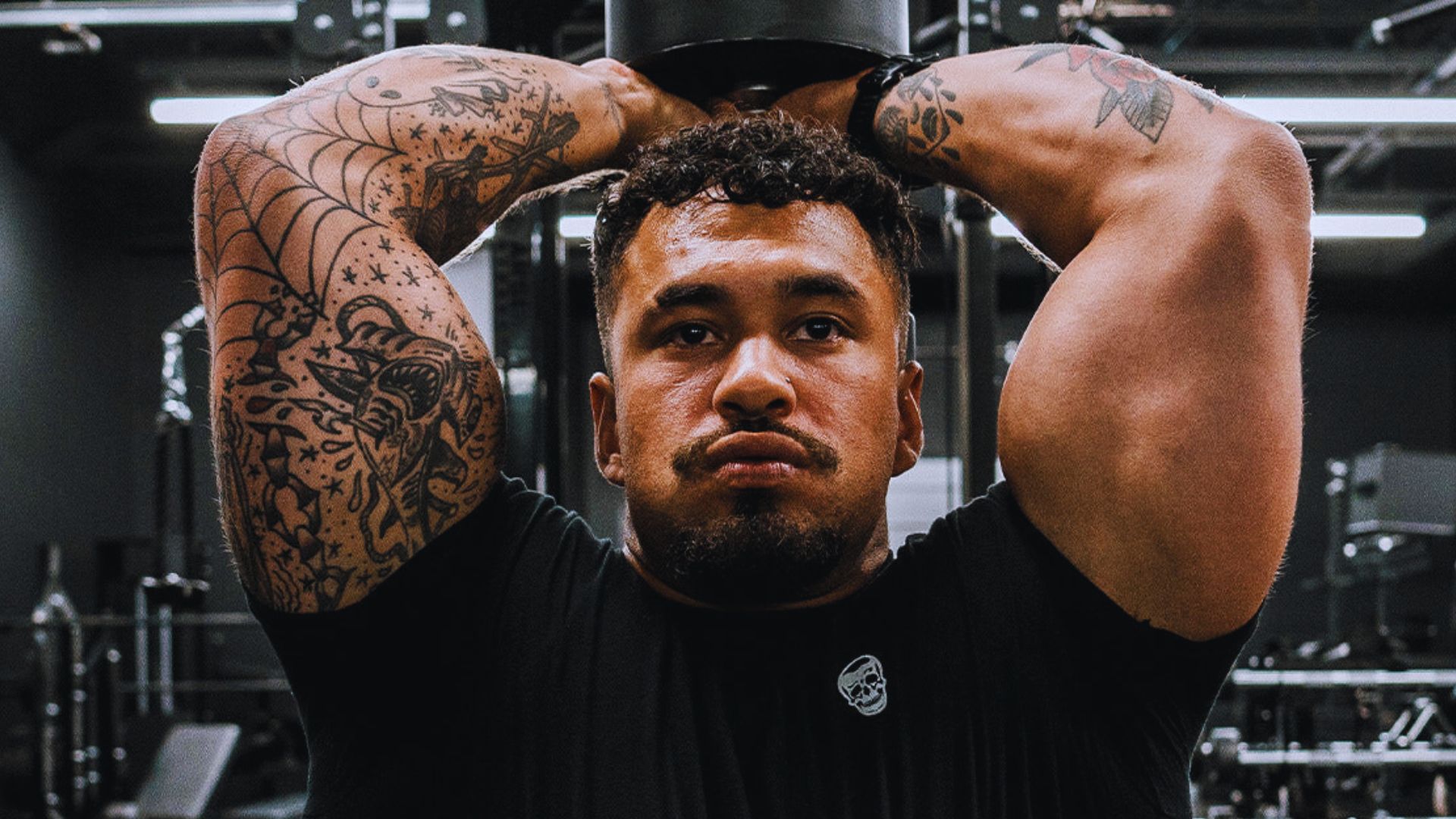 A muscular man with tattooed arms is lifting weights above his head in a gym. His focused expression and the dim setting convey intensity.