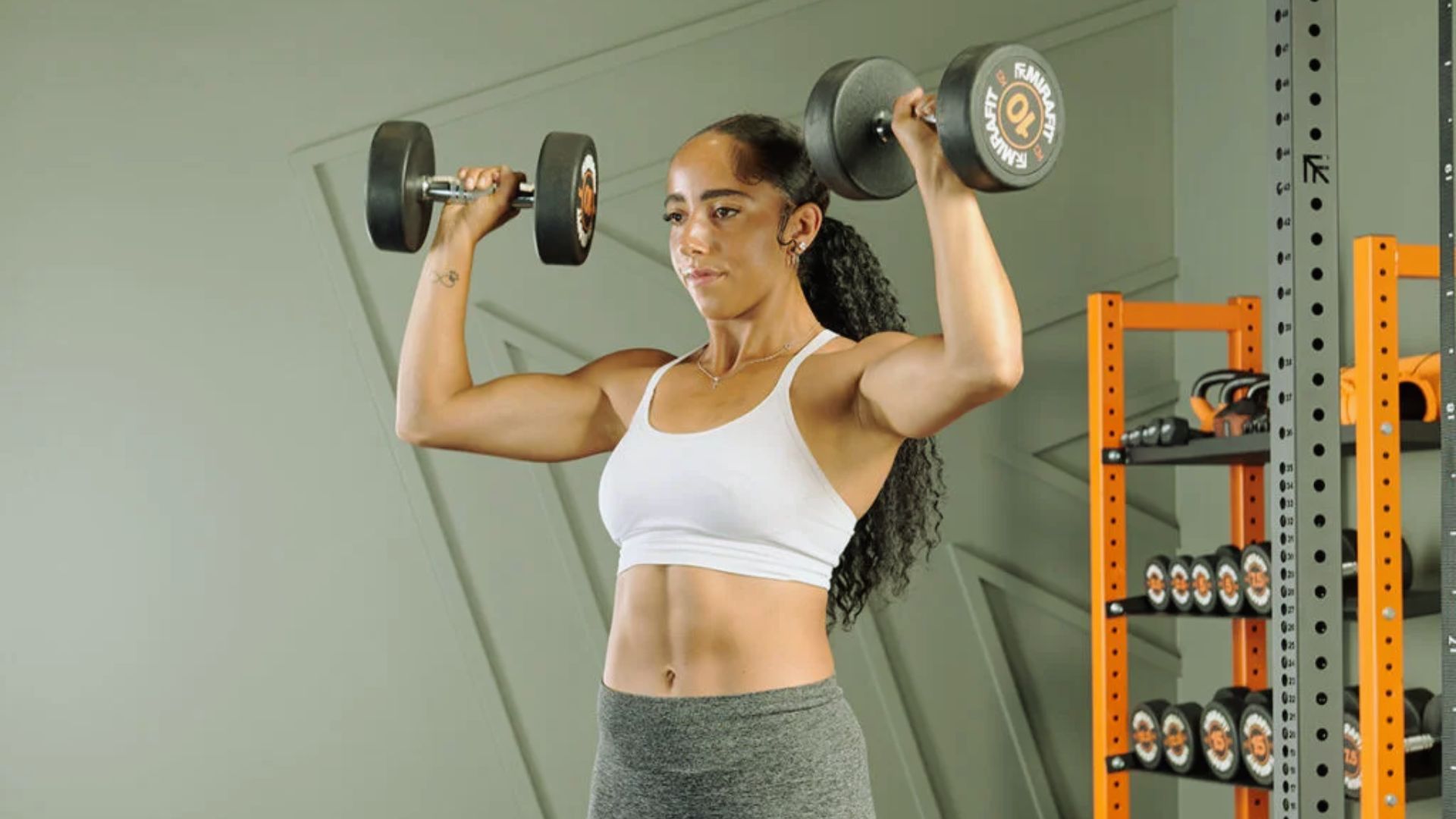 A woman in a white sports bra and gray leggings performs a shoulder press with dumbbells in a gym. She appears focused and strong, with a rack of weights nearby.