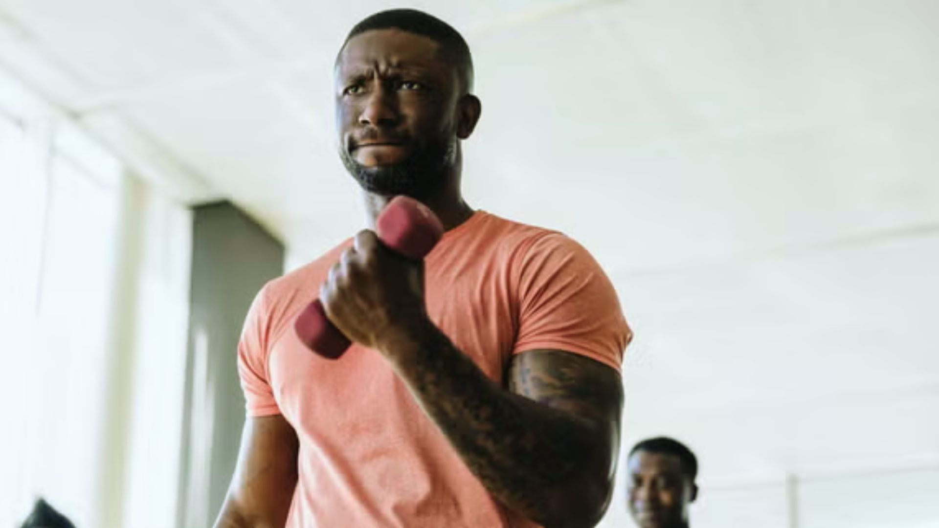 Man focused and determined while lifting a pink dumbbell in a bright gym, wearing a pink shirt. Another person in the background smiles.