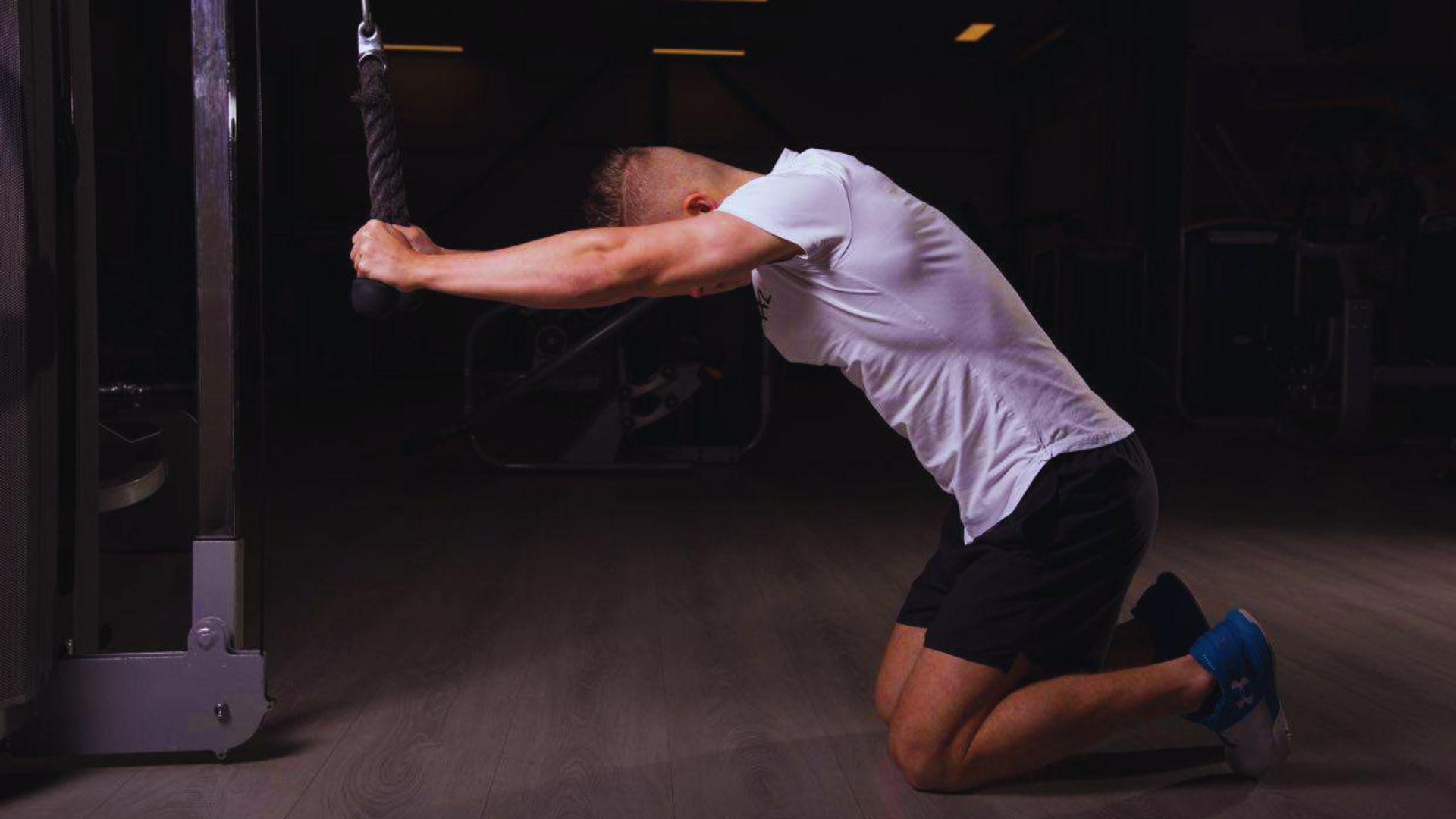 Man kneeling in a gym, wearing a white shirt and black shorts, performing a cable rope exercise. The scene conveys focus and determination.