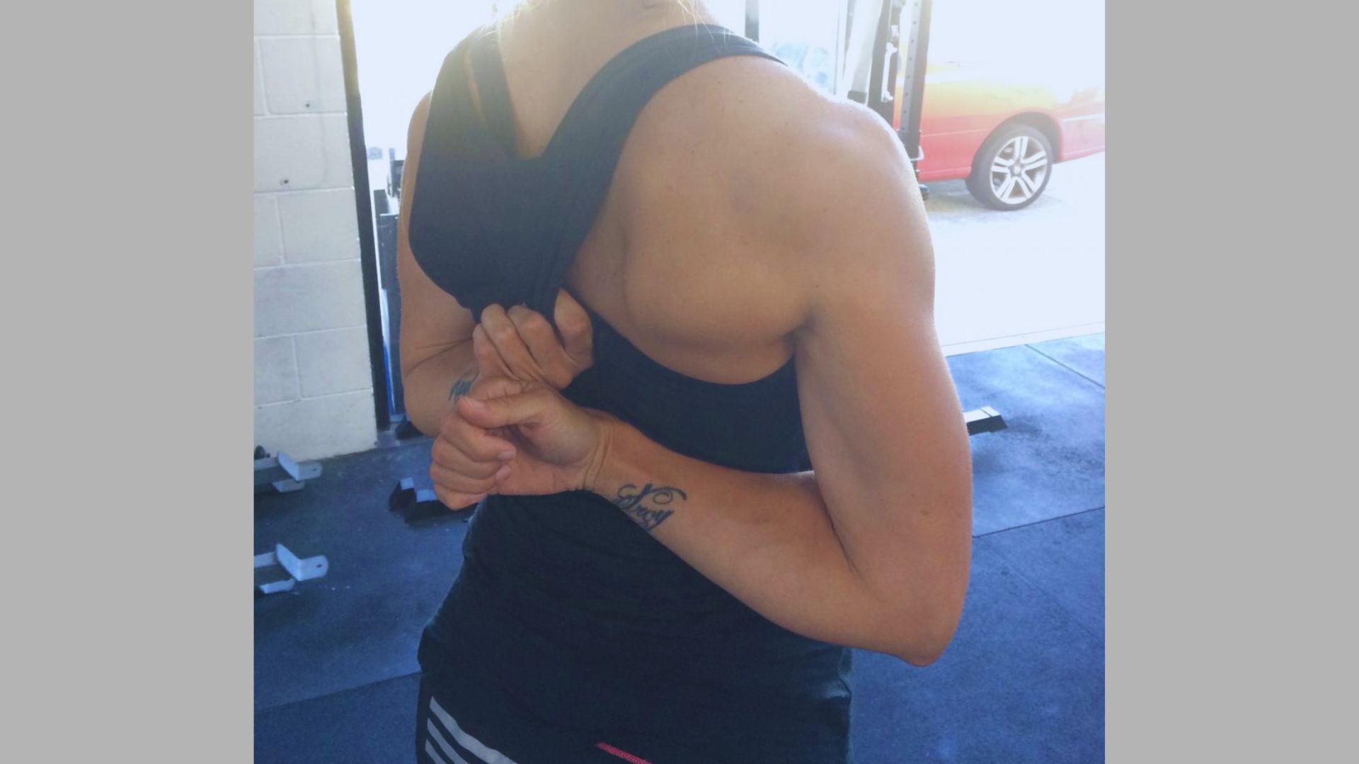 A person in a black tank top does a shoulder stretch in a gym, pulling their arm behind their back. Bright sunlight filters in, highlighting their muscles.