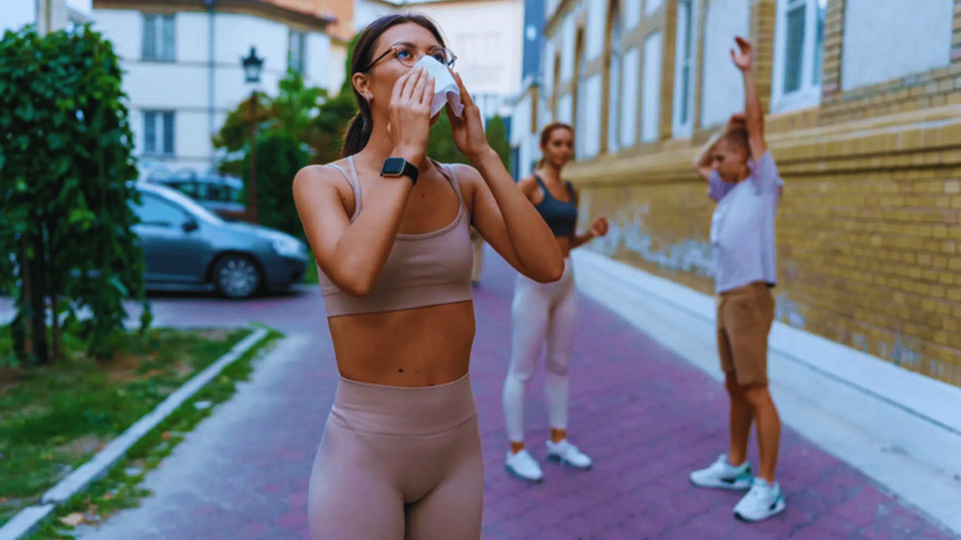 A woman in beige workout clothes wipes her face with a towel outdoors. Two people stretch in the background. The scene conveys a post-exercise relaxation.