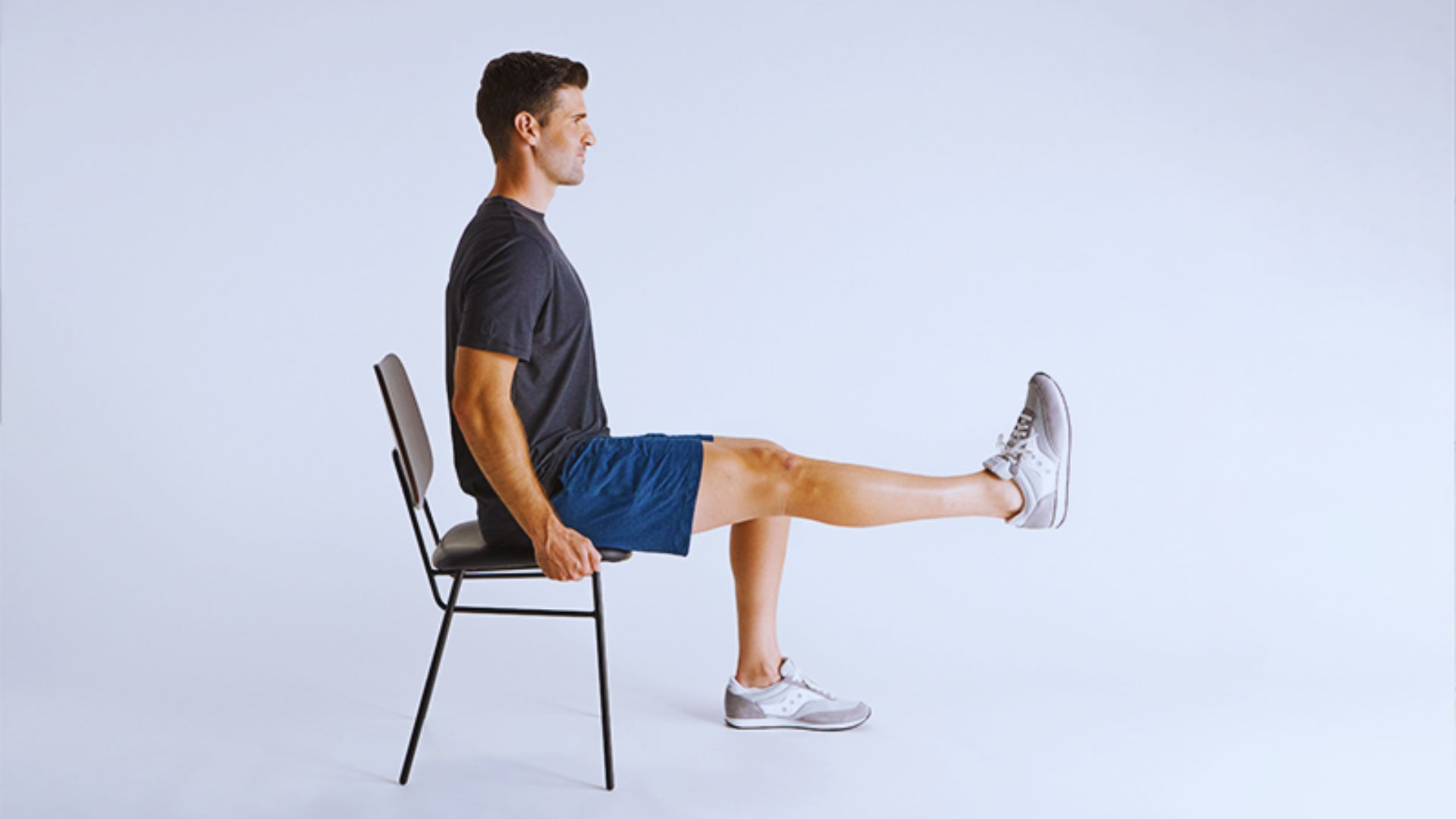 A person in a black shirt and blue shorts sits on a chair, lifting one leg straight. The light background emphasizes a calm, focused exercise moment.