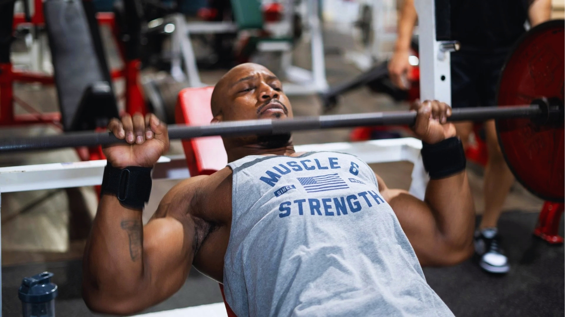 A man intensely focused on bench pressing in a gym, wearing a "Muscle & Strength" tank top. The atmosphere is energetic and determined.