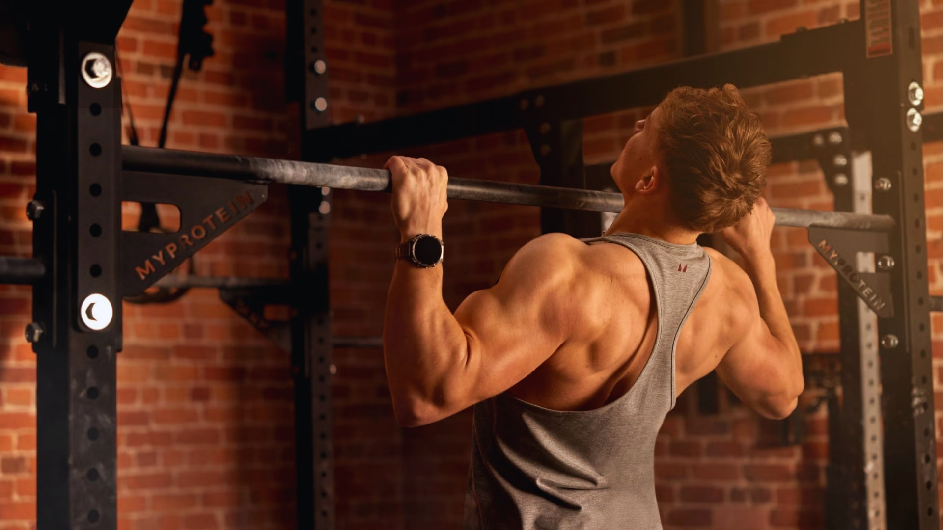 A person doing pull-ups in a gym, wearing a gray tank top and smartwatch. The setting has red brick walls and black metal equipment, conveying strength and focus.