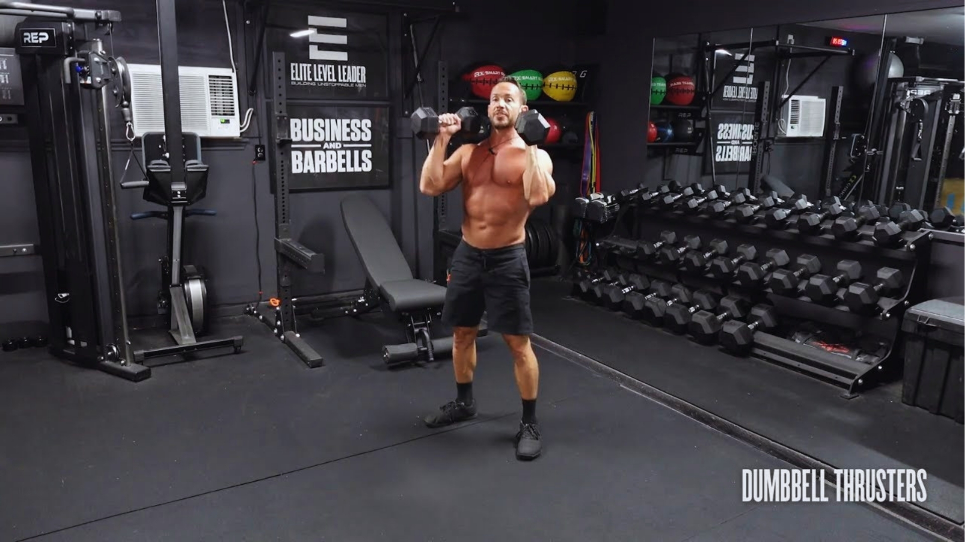 A shirtless man performs dumbbell thrusters in a gym with a rack of dumbbells and medicine balls. The setting is focused and intense.