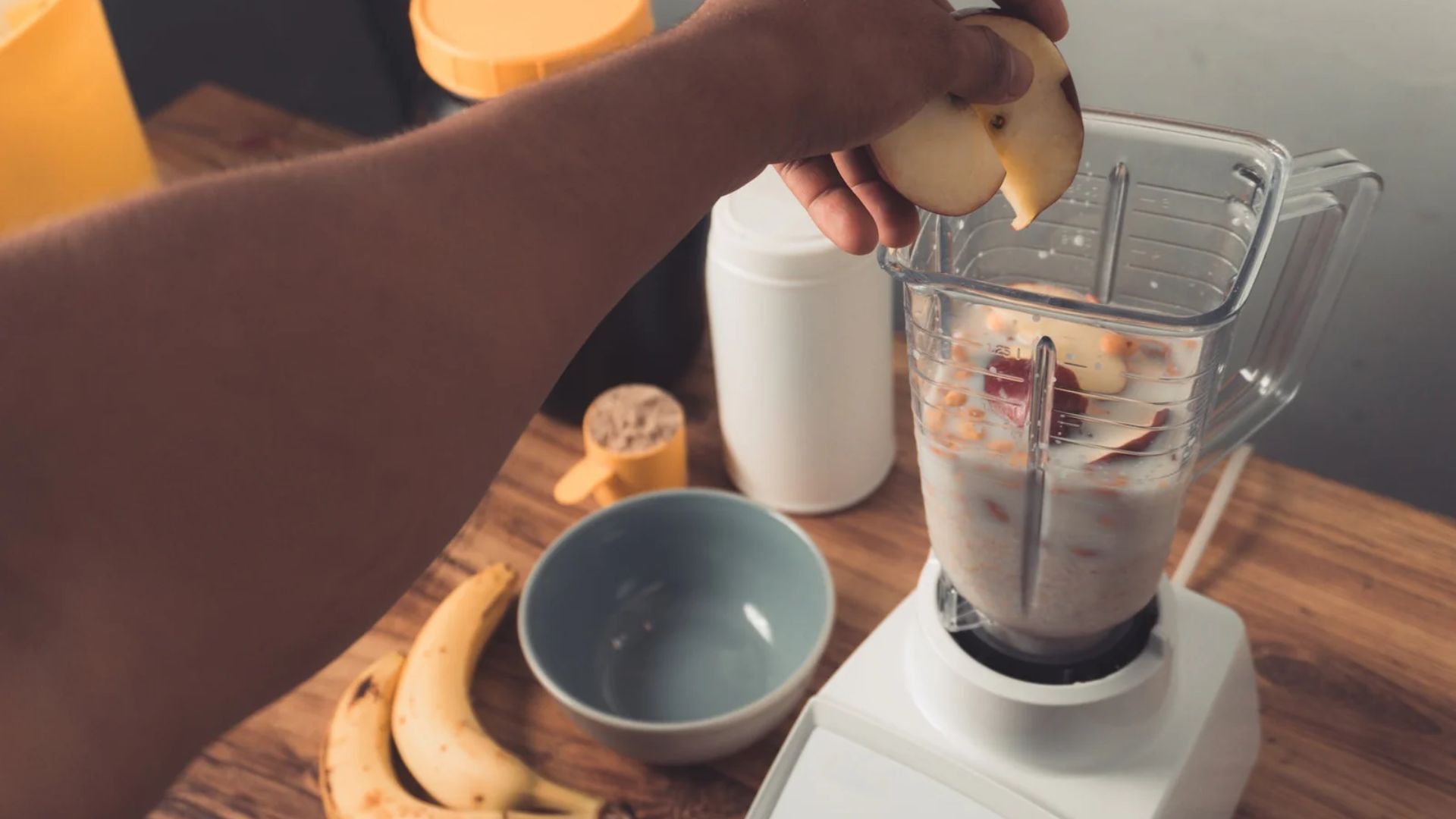 A person blends ingredients in a blender to prepare a smoothie in a bright kitchen setting.