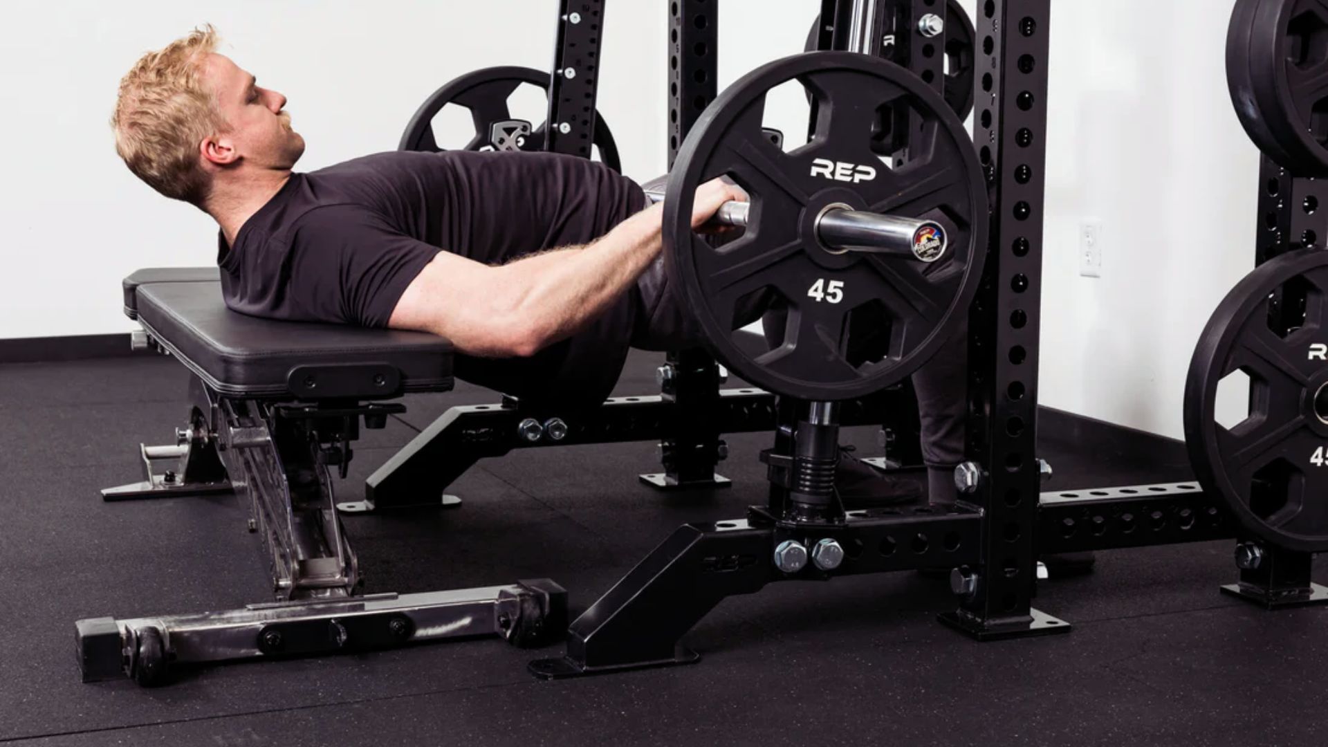 A person performs a hip thrust exercise on a bench with a barbell, concentrating intensely. The gym setting is clean and modern, conveying strength and focus.