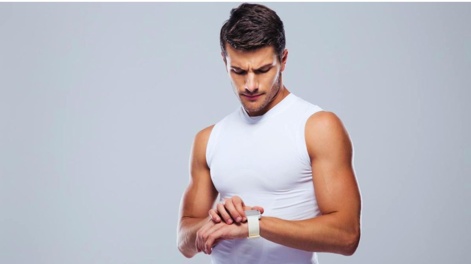A focused man in a sleeveless white shirt looks at his smartwatch. His expression is concentrated, and the neutral background enhances his athletic build.