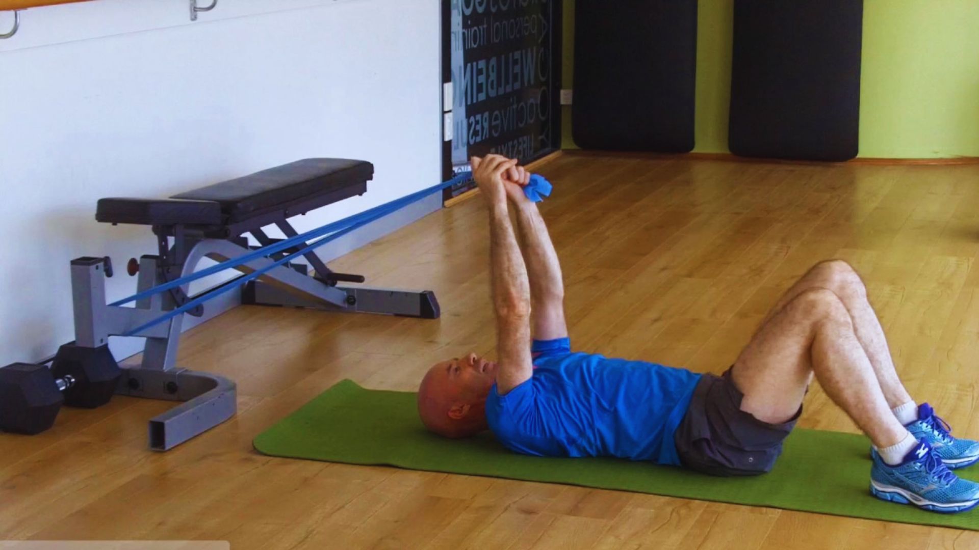 A man in a blue shirt exercises on a green mat, using blue resistance bands attached to a bench. The room has wooden floors, conveying a gym setting.