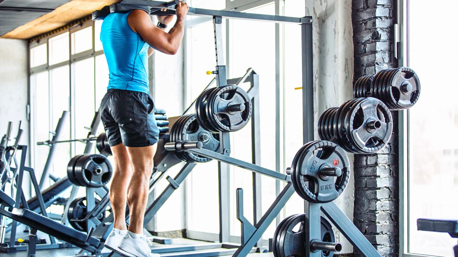 A man in a blue tank top and black shorts is doing pull-ups in a gym. He is surrounded by weightlifting equipment with natural light streaming through large windows.