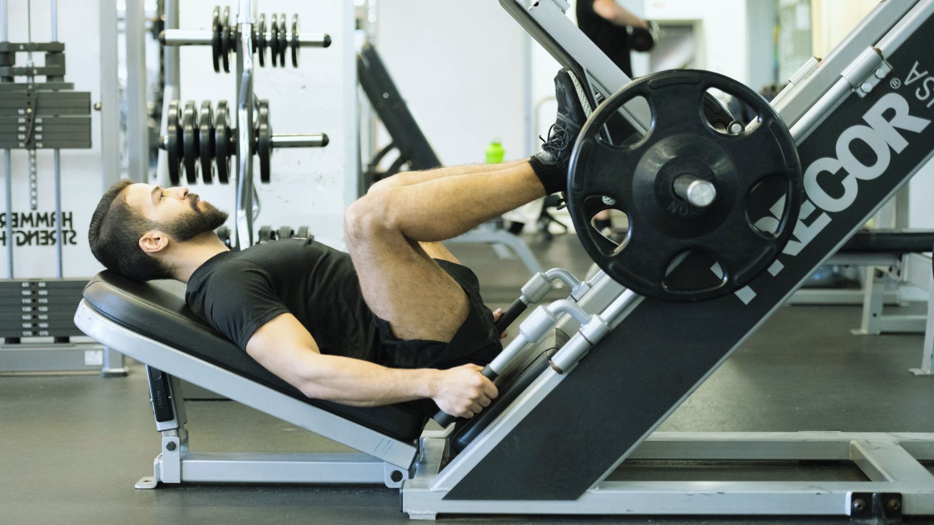 A person with a beard is using a leg press machine in a gym. They are wearing a black shirt and shorts. The setting is organized, with weights visible.