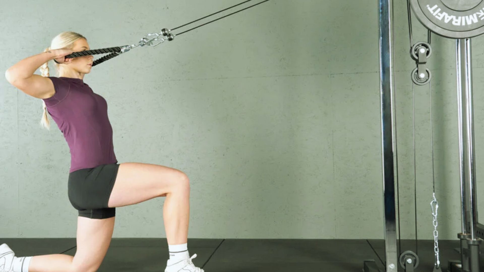 A person performs a face pull exercise on a cable machine, kneeling on one knee. They wear a purple shirt and black shorts, focused and determined.