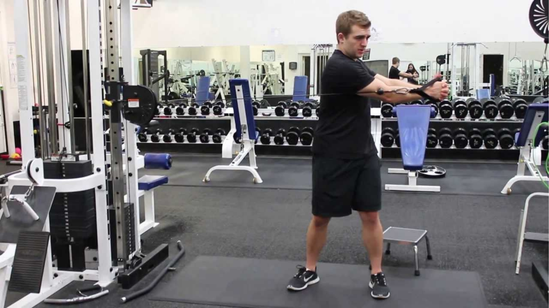 A man in a gym performs a cable machine exercise, focused and engaged. He's wearing a black shirt and shorts, surrounded by gym equipment and mirrors.