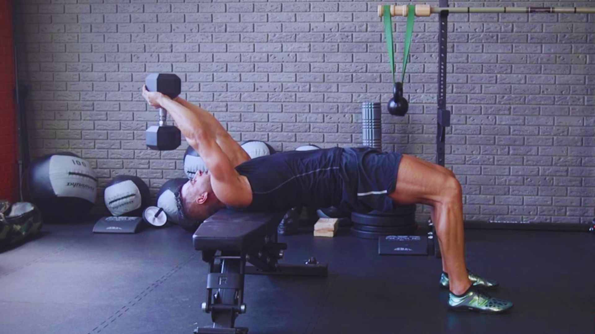 Man performing a dumbbell pullover on a bench in a gym. He is in profile view, wearing athletic attire, with exercise equipment visible around him.