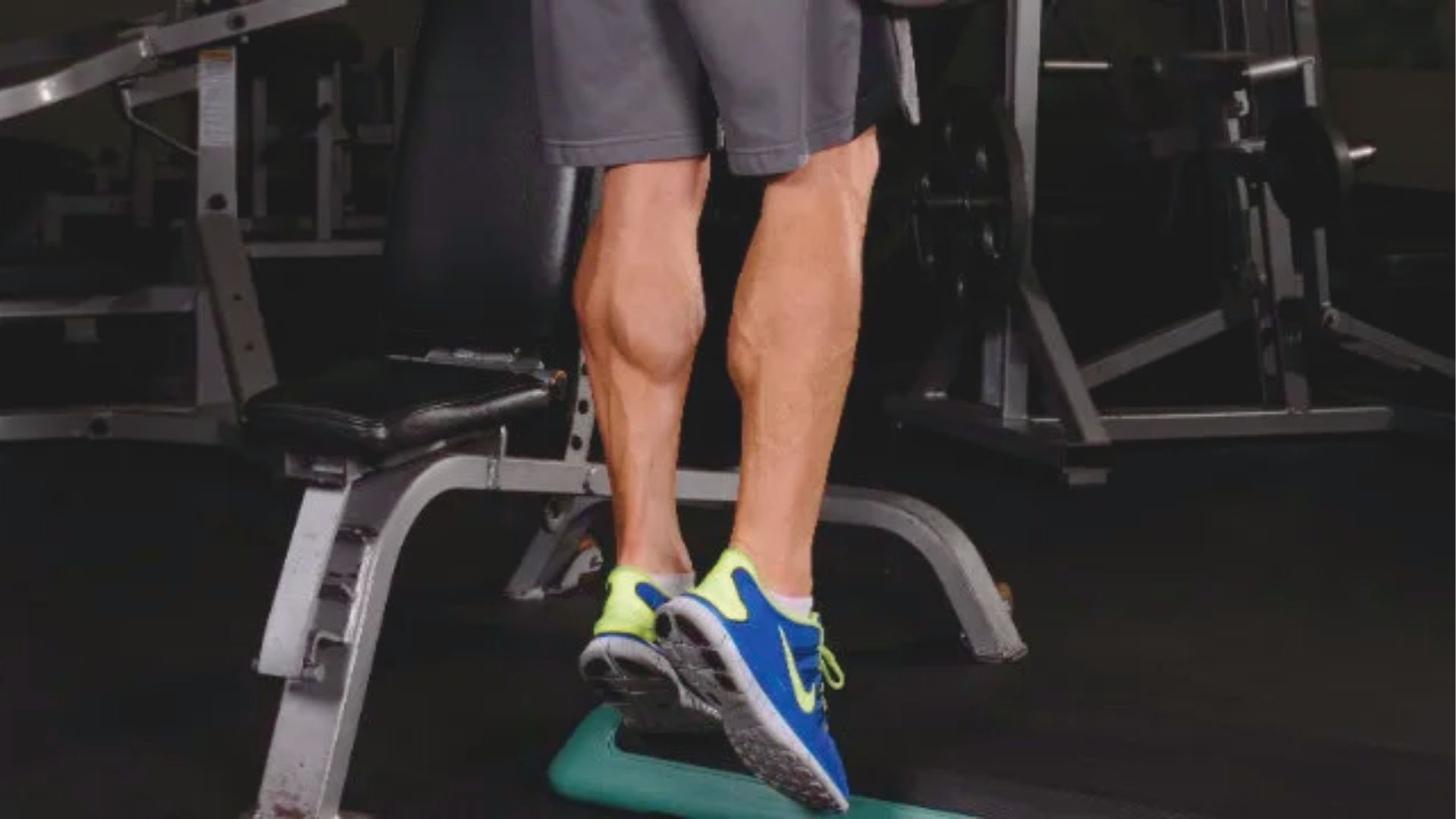 A person in sneakers performs a toe raise exercise at a gym. Their calf muscles are flexed, and various gym equipment is visible in the dim background.