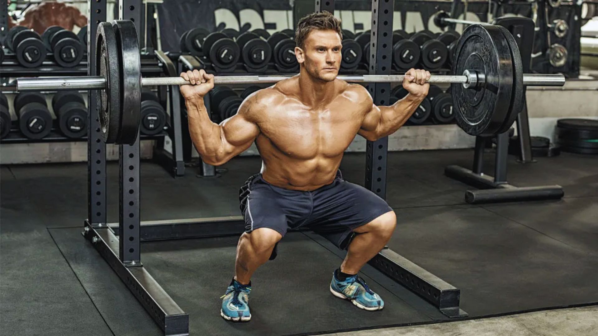 A muscular man performs a barbell squat in a gym, focused and determined. He's shirtless, wearing shorts and sneakers, with weights and equipment visible.
