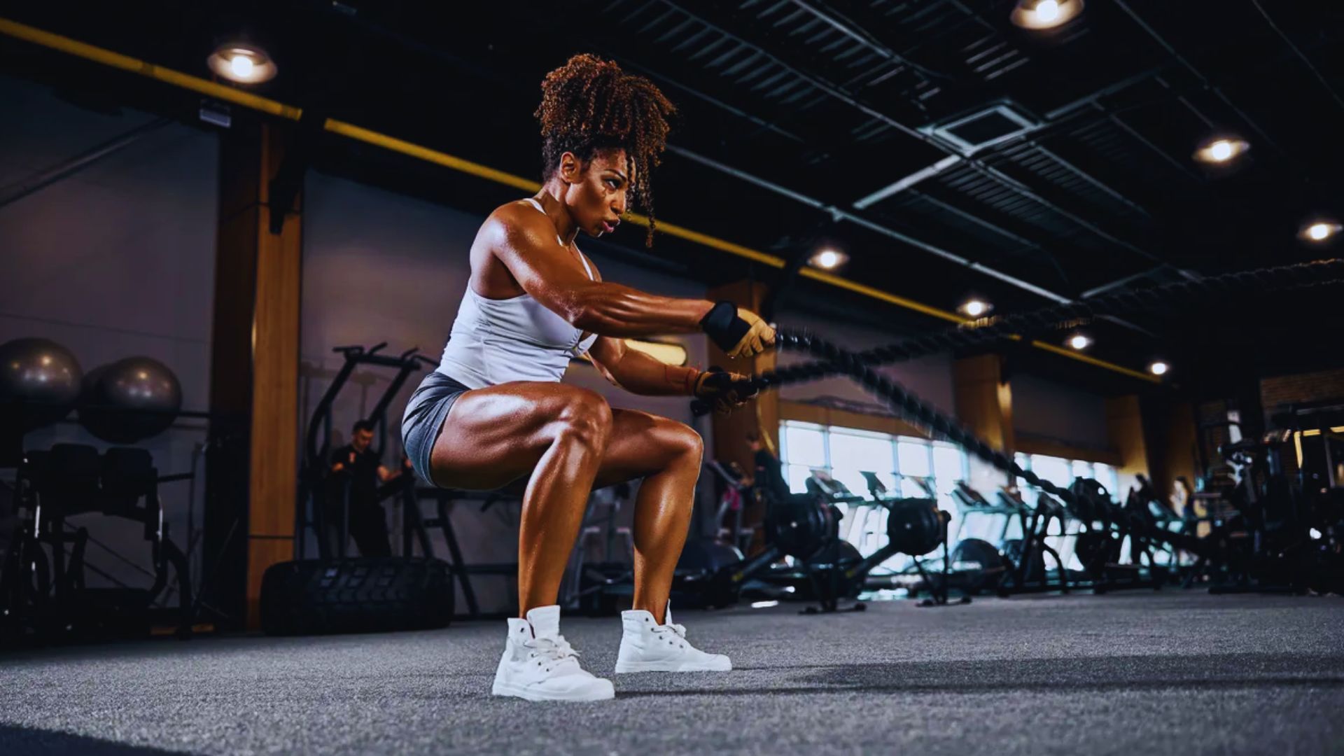 A determined woman in athletic wear vigorously exercises with battle ropes in a well-equipped gym, showcasing strength and focus under bright lights.