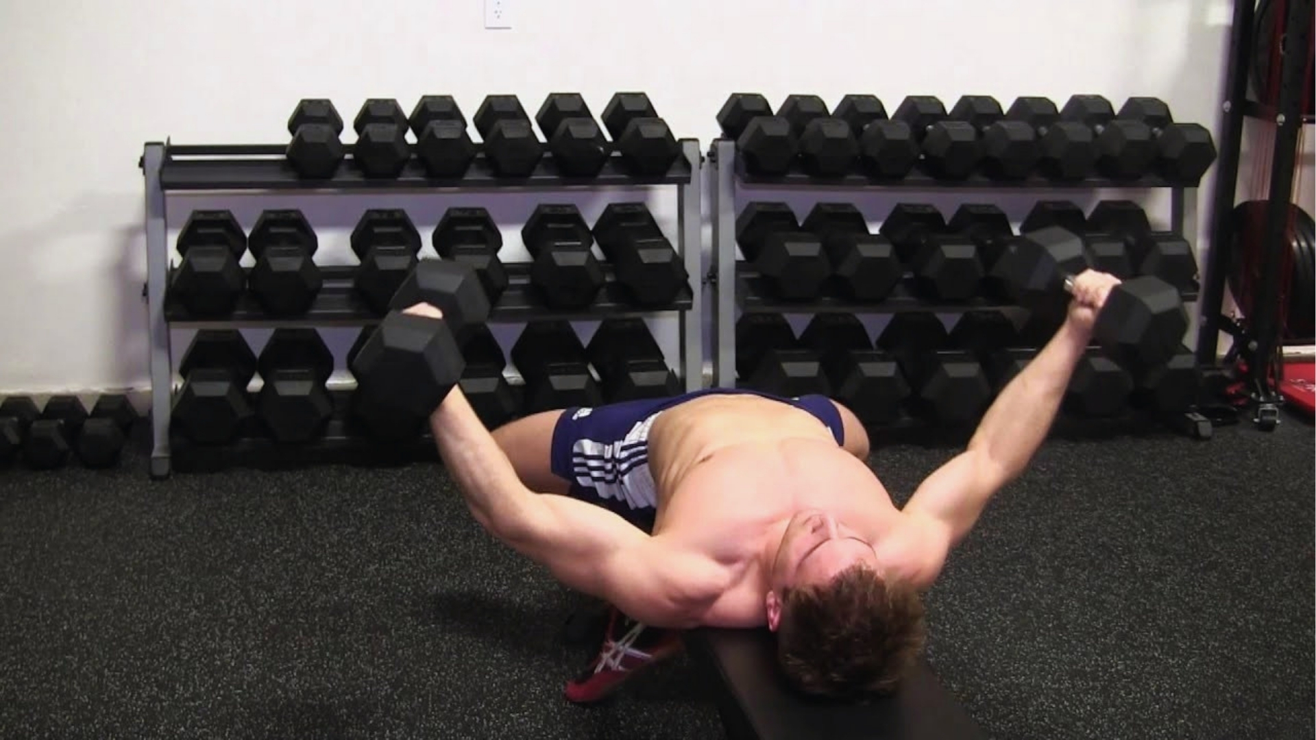 A man performs a dumbbell chest press on a bench in a gym, surrounded by a rack of dumbbells. The setting is focused and determined.