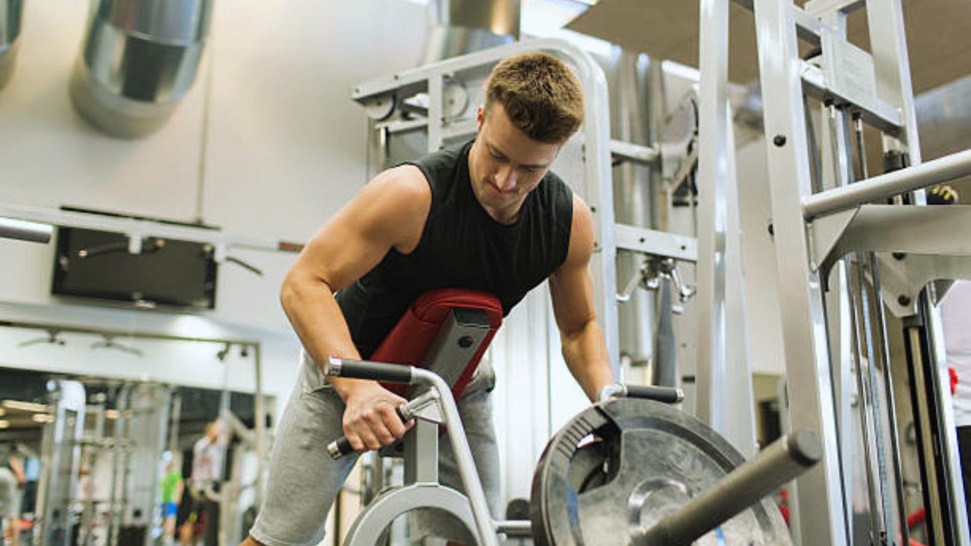 Man in a gym using a rowing machine, wearing a black sleeveless shirt and gray shorts. He appears focused and determined amidst exercise equipment.