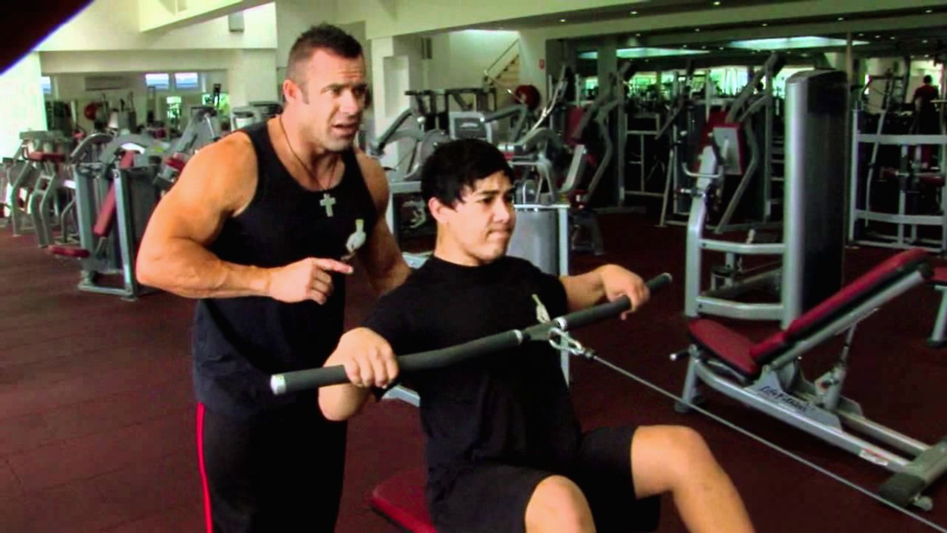 A muscular trainer coaches a young man on a rowing machine in a gym. The setting is energetic, with exercise equipment visible in the background.