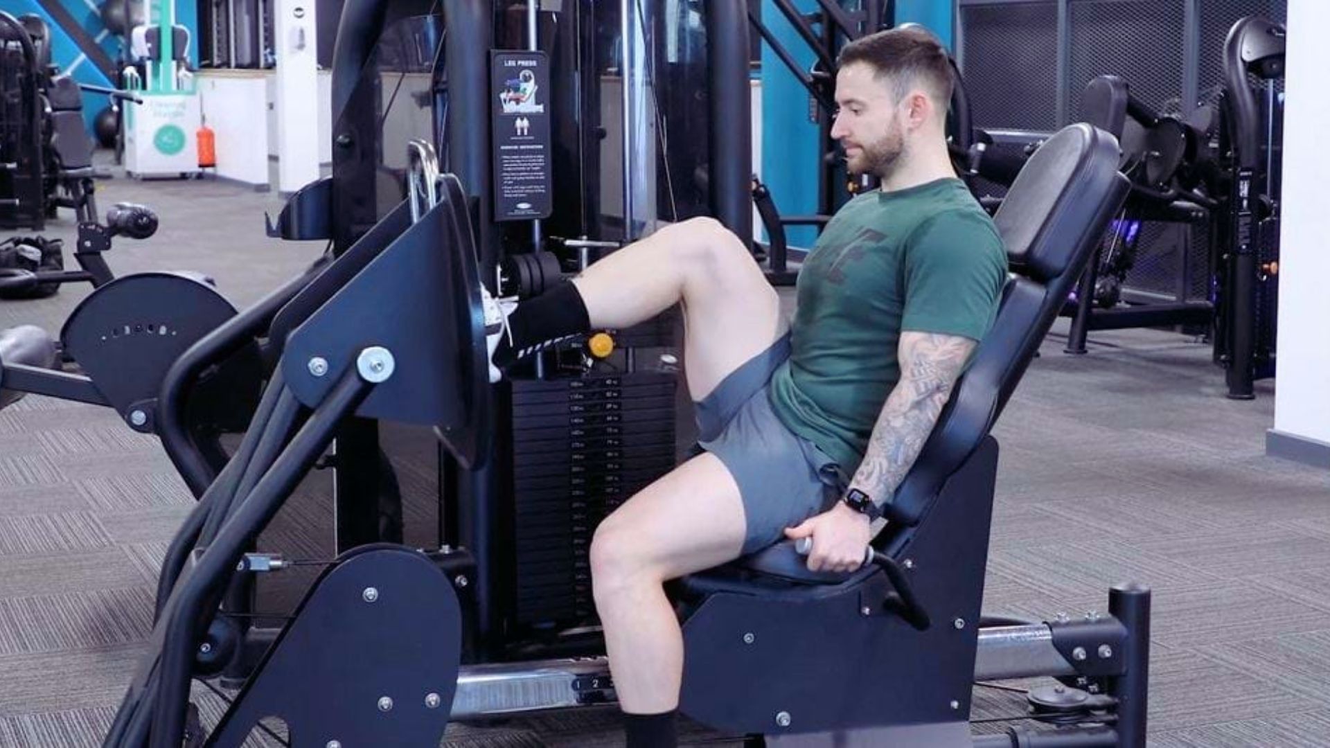A person in athletic wear uses a leg press machine at the gym, focusing intently. The setting is modern, with equipment and weights visible around.