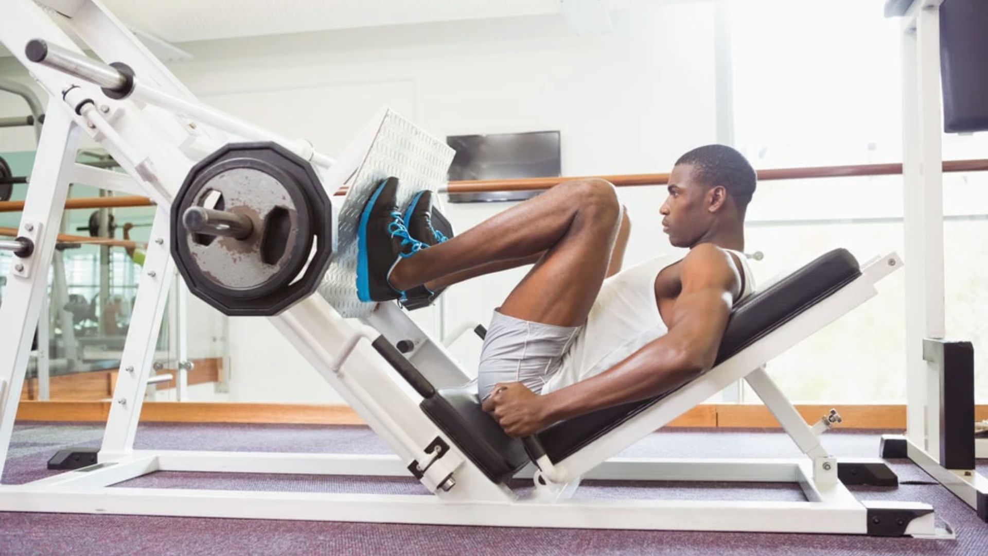A person is using a leg press machine in a gym, wearing a white tank top and gray shorts. They appear focused and determined, performing an exercise.