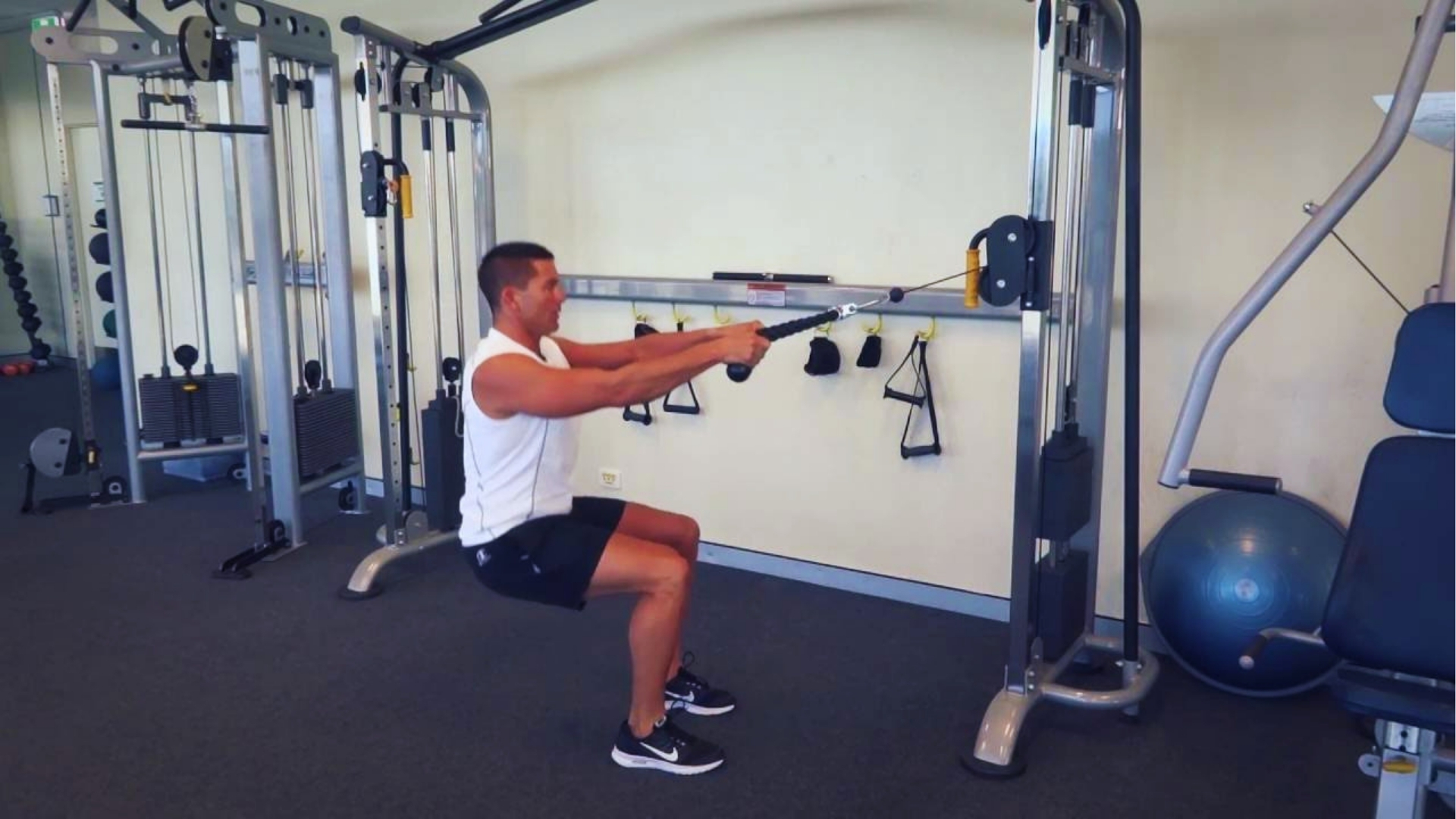 A man in a white tank top and black shorts uses a cable machine for squats in a gym. He is focused and maintaining good form. Fitness equipment surrounds him.
