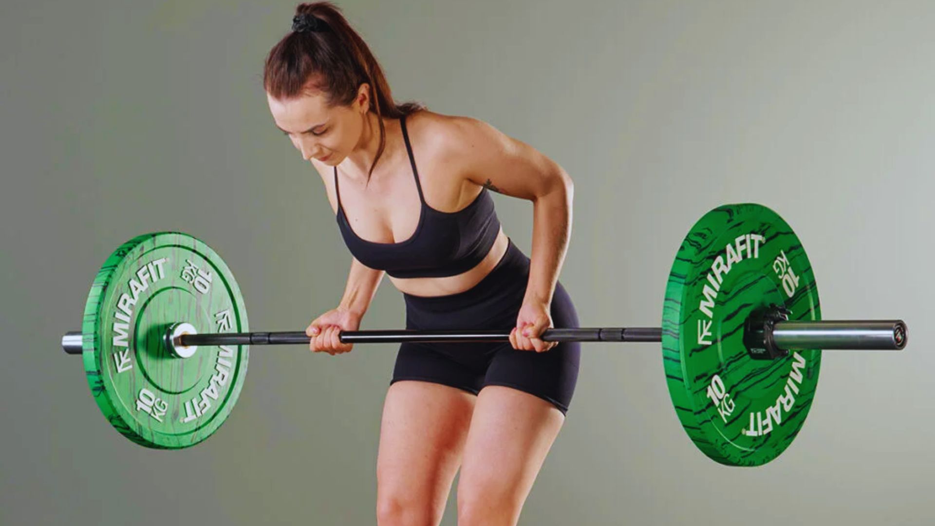 A woman in a black sports outfit performs a bent-over row with a barbell. Green weight plates are on each side. She looks focused and strong.