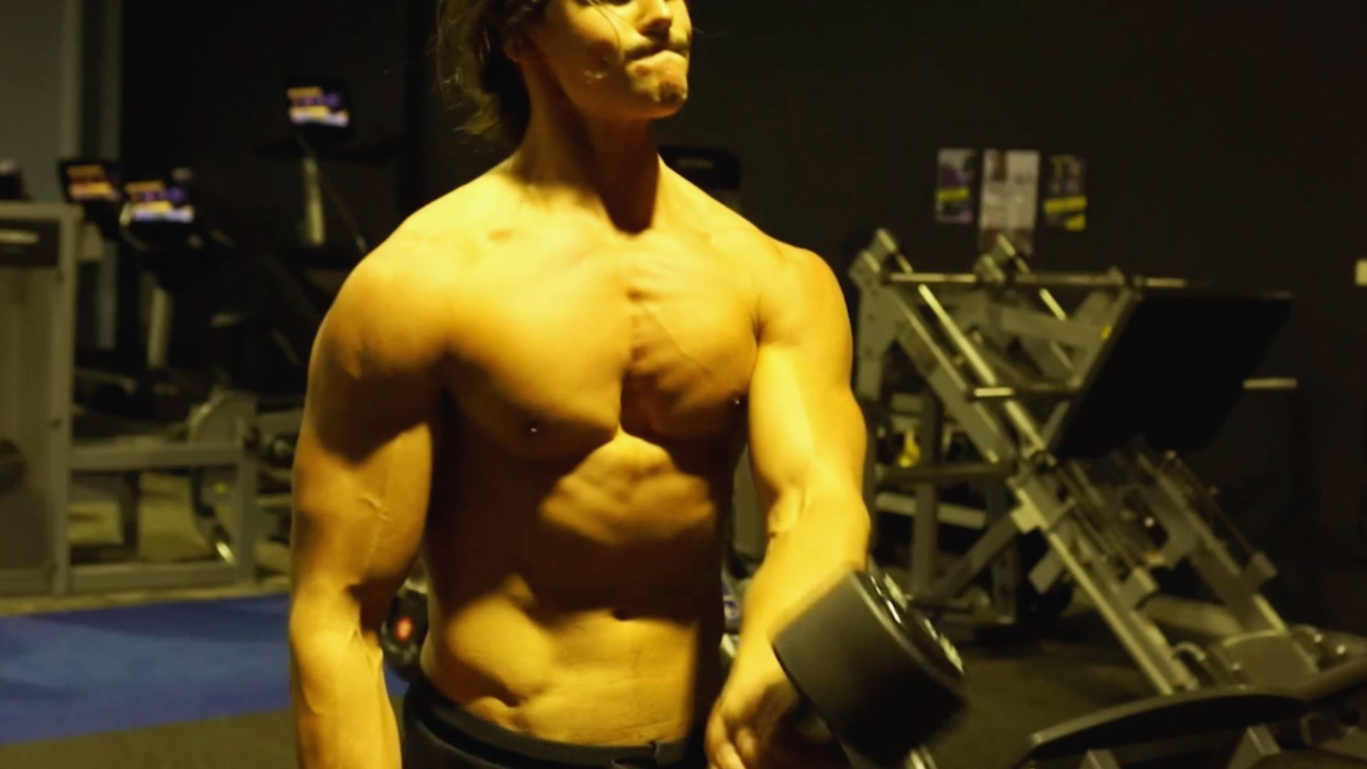 A muscular person lifts a dumbbell in a dimly lit gym. The focused expression suggests determination. Exercise equipment is visible in the background.
