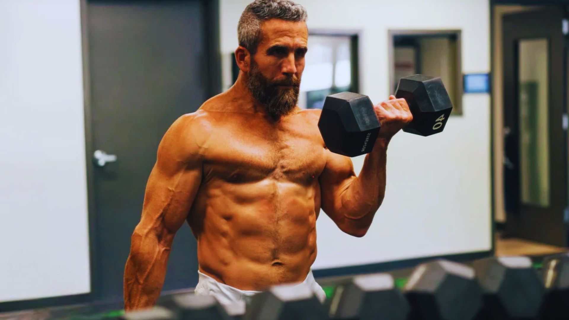 A muscular man with a beard performs a bicep curl with a 40-pound dumbbell in a gym, showcasing strength and focus. Gym equipment is in the background.