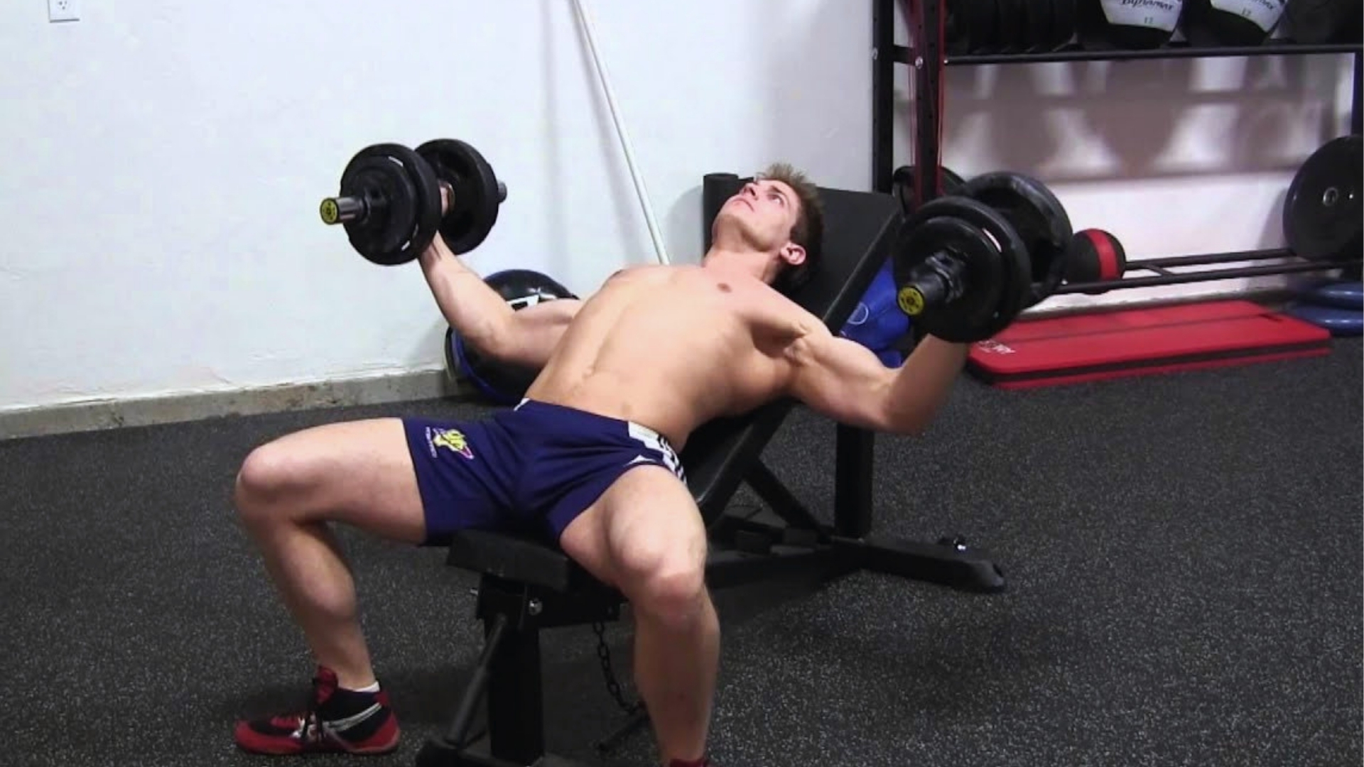 A person performs an incline dumbbell chest press on a bench in a gym. They are shirtless, wearing shorts, and appear focused, surrounded by gym equipment.