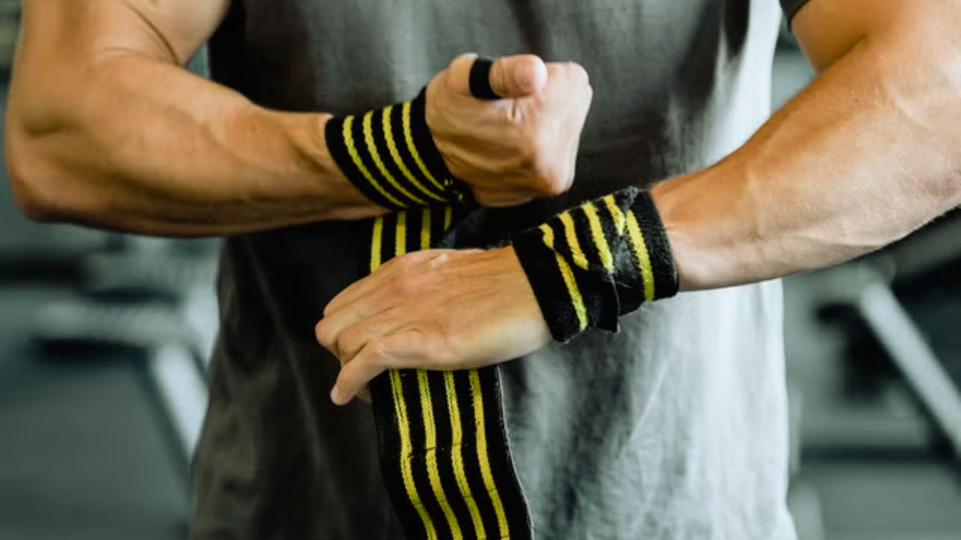 A person wraps a striped black and yellow wrist wrap around their wrist in a gym setting, conveying preparation and focus on strength training.