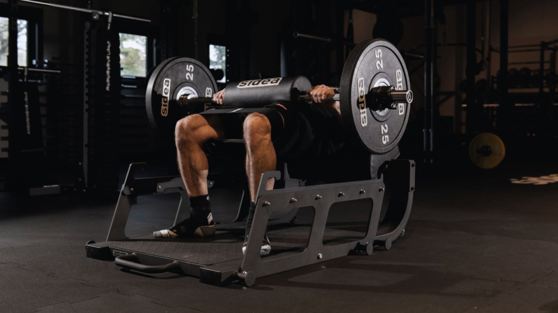 Man performing a barbell bench press on a hip thrust machine in a dimly lit gym. Plates on the bar read "25" with the brand "Sidea" visible.