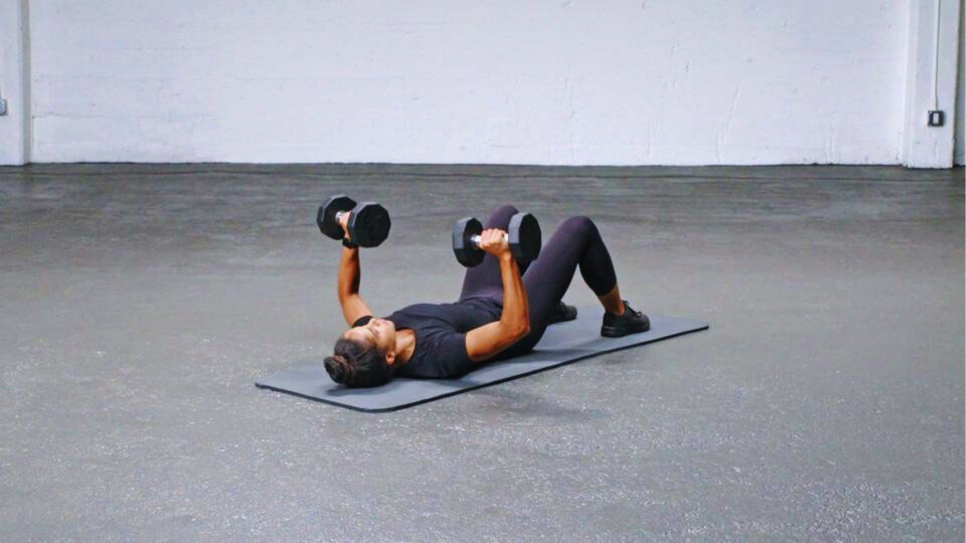 A person in athletic attire performs a dumbbell chest press on a gym mat in a minimalist room. The focused expression conveys strength and determination.