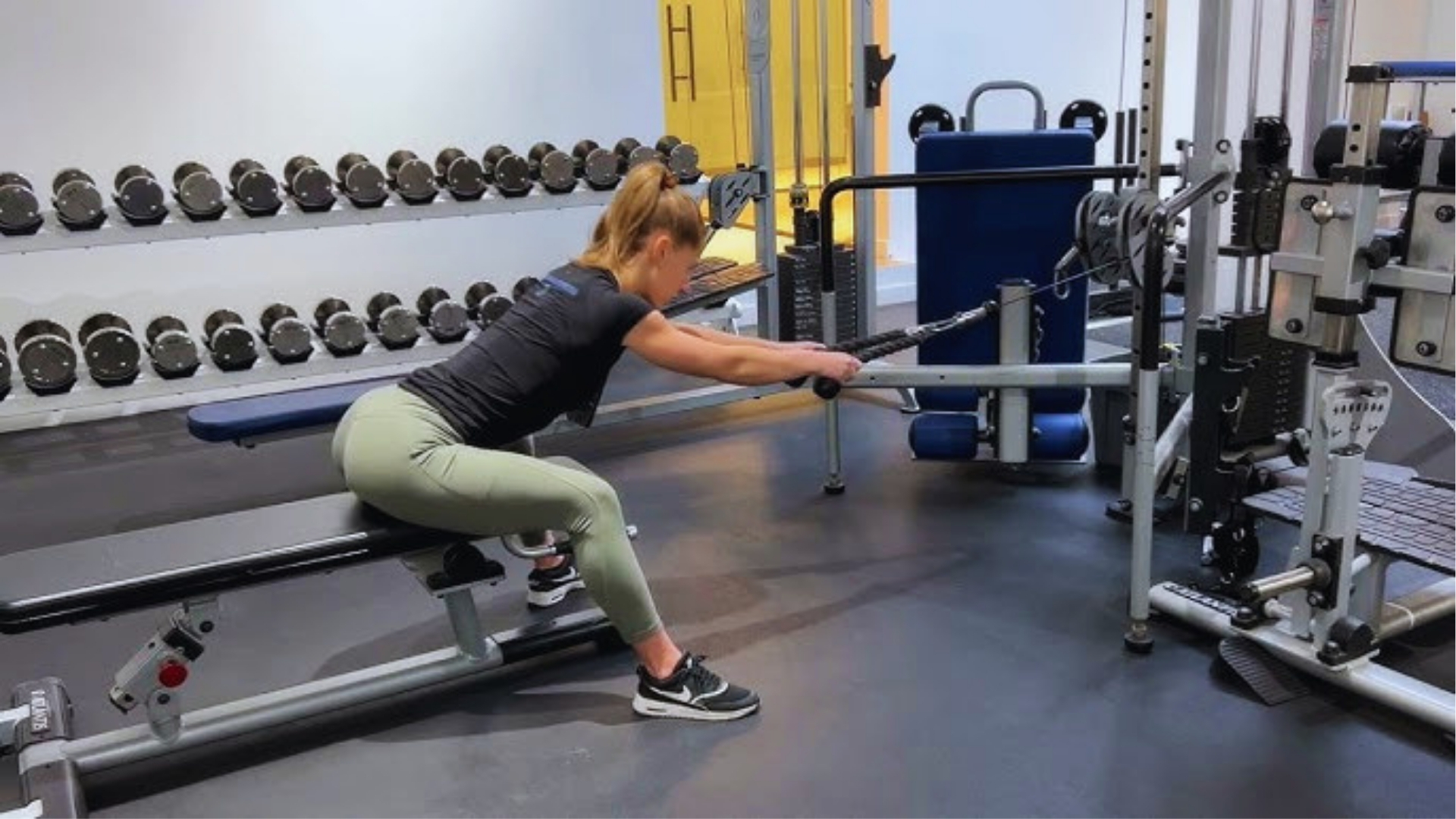 Woman in a gym doing a seated cable row exercise on a machine. She's focused, wearing a black top and green leggings. Dumbbells are visible in the background.