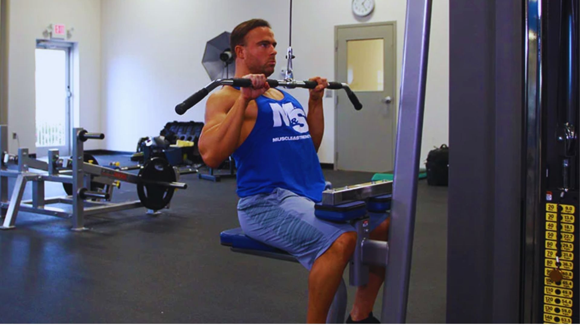A man in a blue tank top uses a lat pulldown machine in a gym, focused and determined. Exercise equipment and a clock are visible in the background.