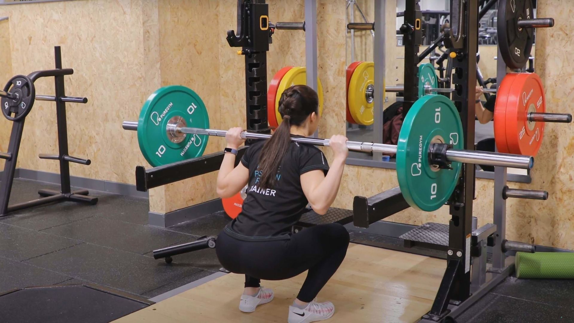 A woman performs a barbell squat on a platform in a gym, surrounded by weightlifting equipment. She wears workout clothes, conveying focus and strength.