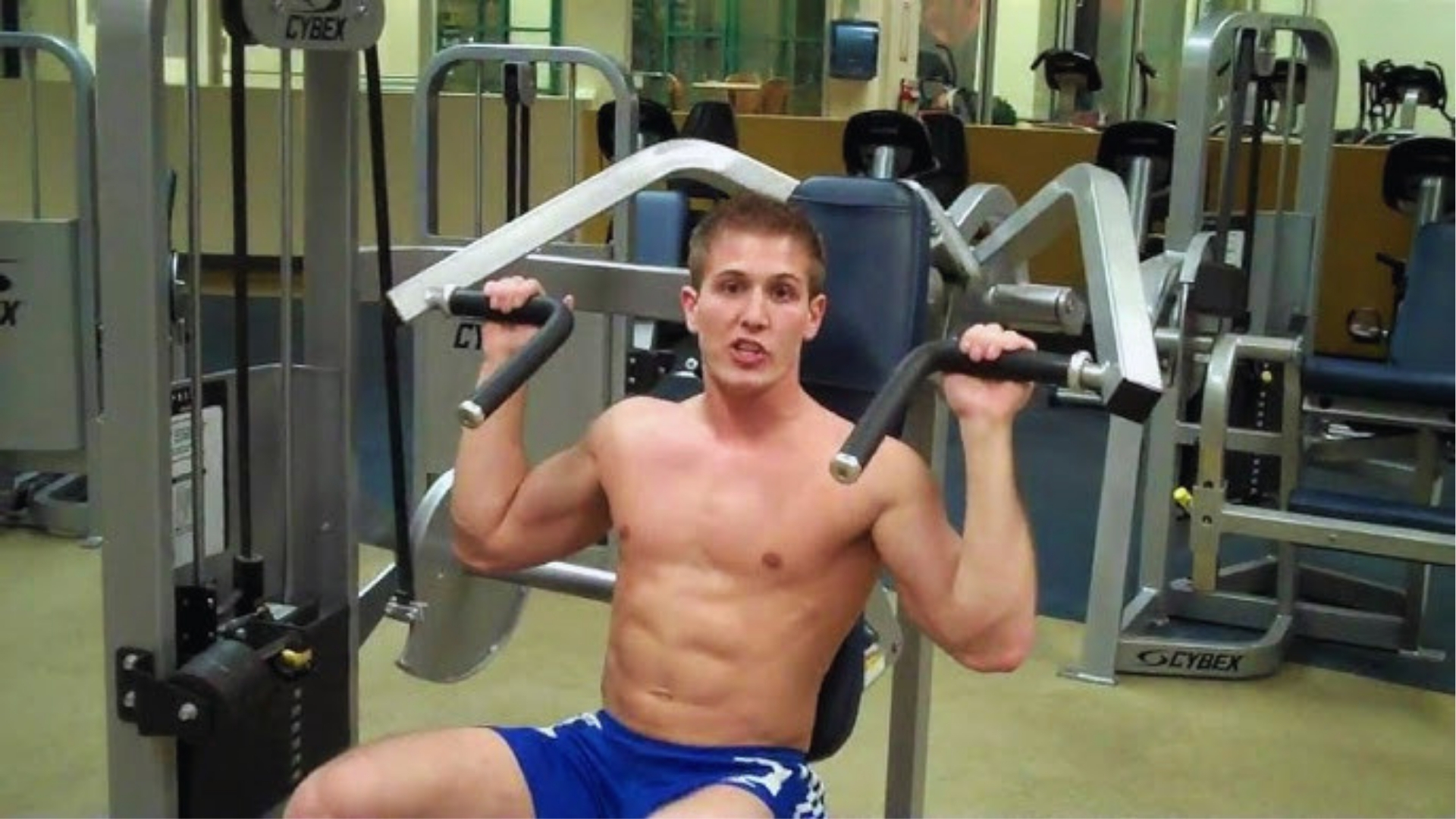 A man is seated on a Cybex machine in a gym, performing an overhead press. He is shirtless, wearing blue shorts, and appears focused and determined.