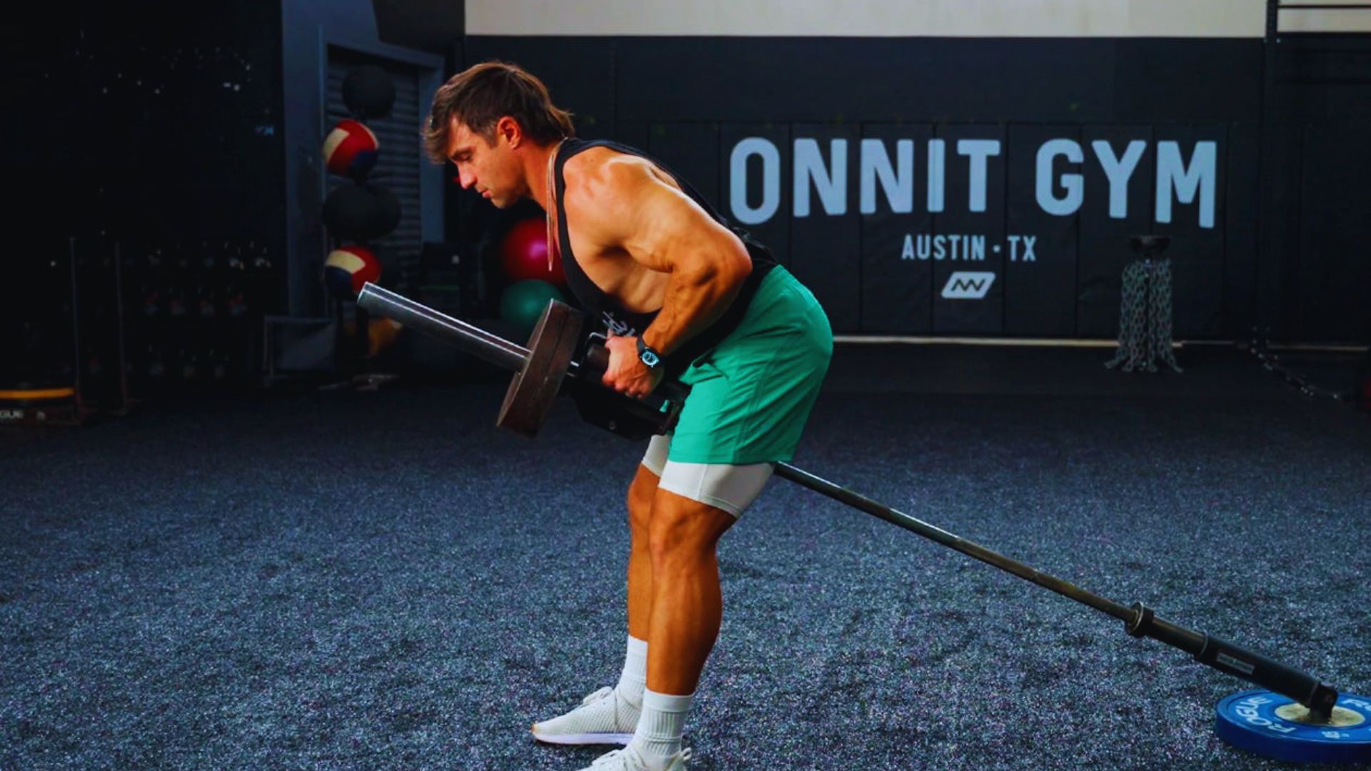 A man in a gym performs a bent-over barbell row with focus. He's wearing a black tank top and teal shorts. The gym wall reads "Onnit Gym, Austin TX."