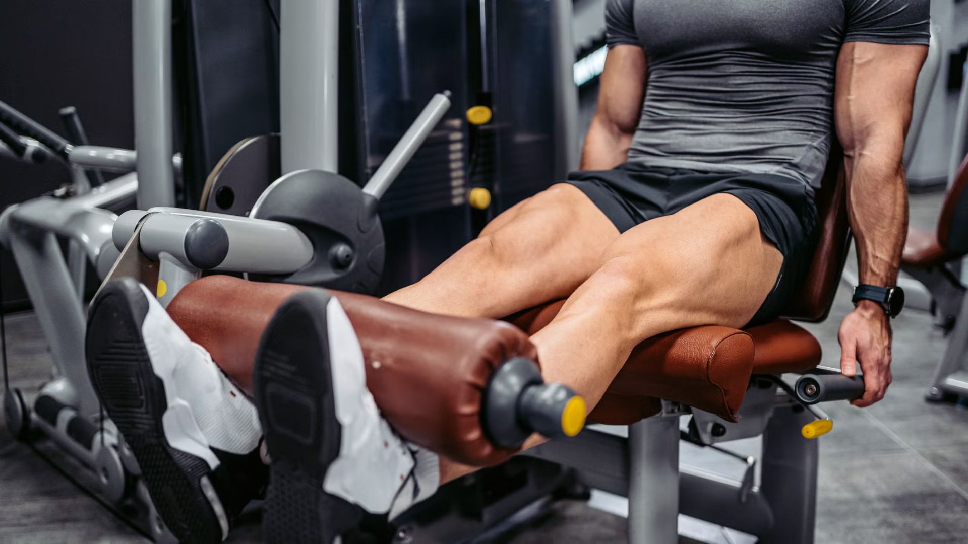 A man in a gym uses a leg extension machine, emphasizing his muscular legs. He wears a gray shirt and black shorts, focusing intensely on his workout.