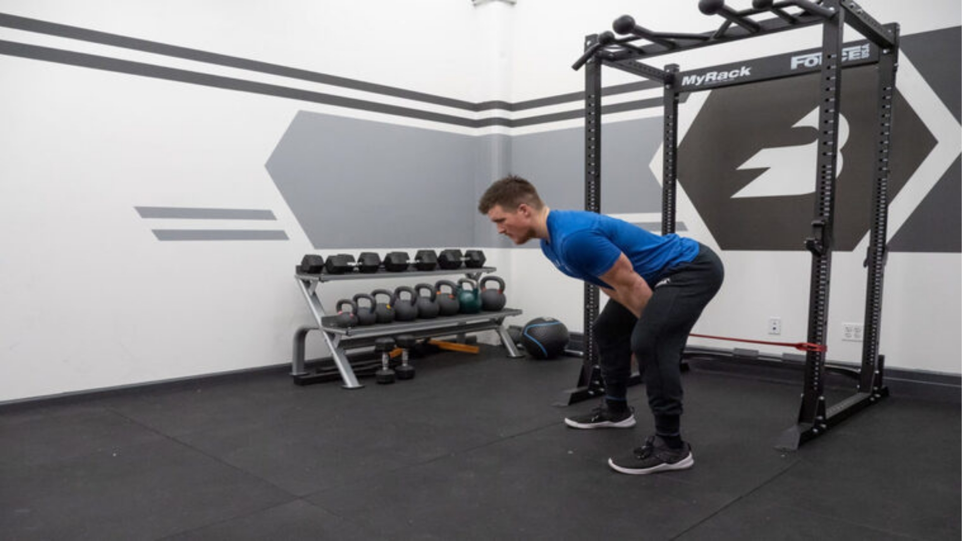 A person in a blue shirt performs a bent-over row in a gym. Behind them, a rack with weights and kettlebells lines the wall, creating an active, focused atmosphere.