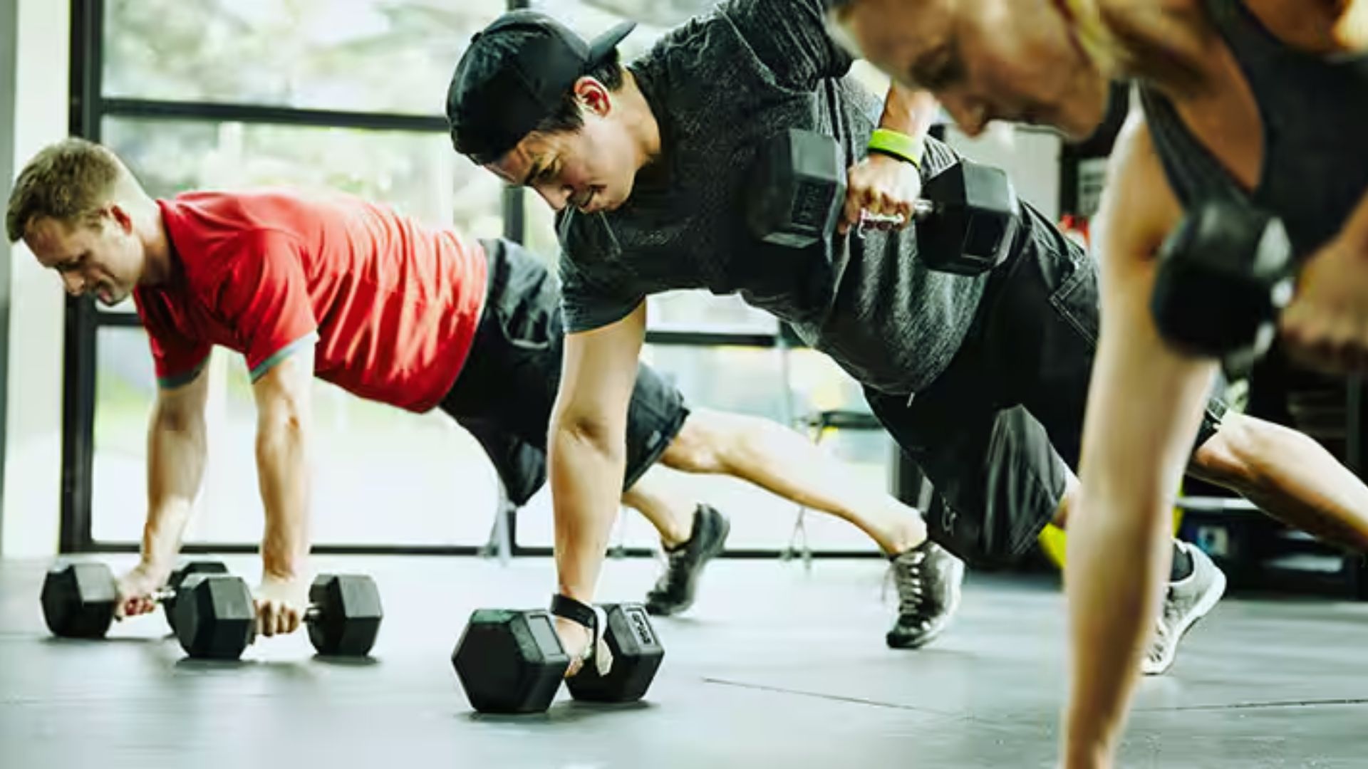 Three people in athletic wear are performing push-ups using dumbbells in a gym. They appear focused and determined, with natural light in the background.