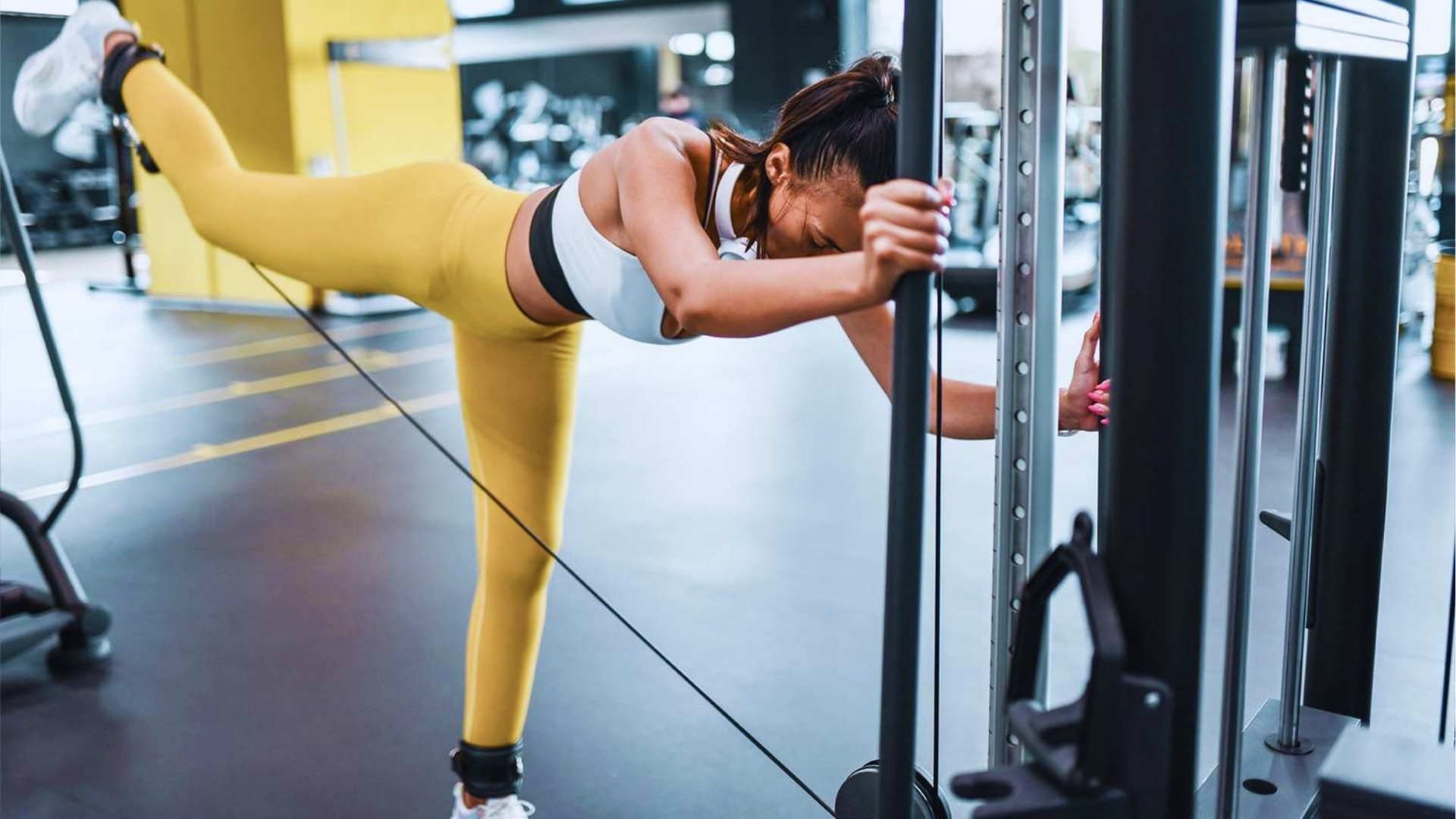 A woman in yellow leggings and a white sports bra exercises on a pulley machine at the gym, performing a leg extension with focus and determination.