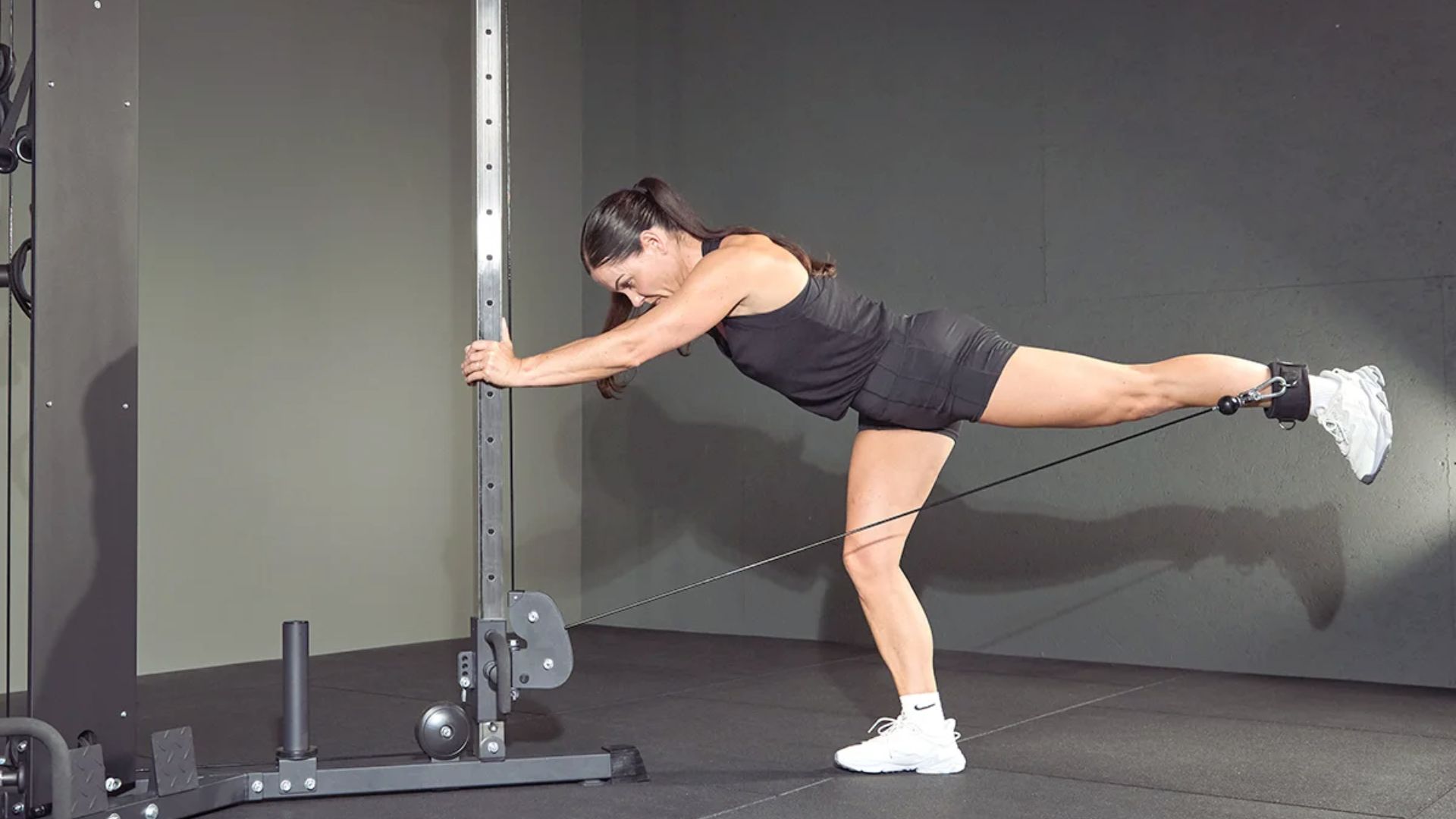 A woman in athletic wear performs a single-leg cable kickback at the gym, poised and focused, highlighting strength and balance against a neutral background.