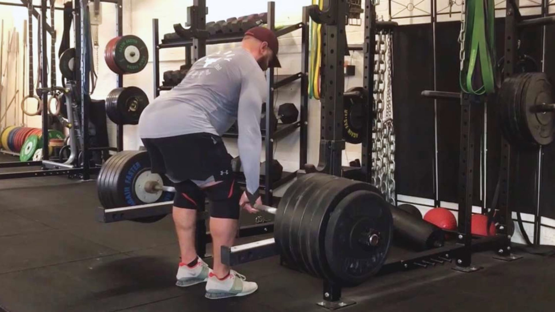 A man in a gym is performing a deadlift with heavy weights. He's wearing a cap, long-sleeve shirt, and shorts, surrounded by weightlifting equipment.