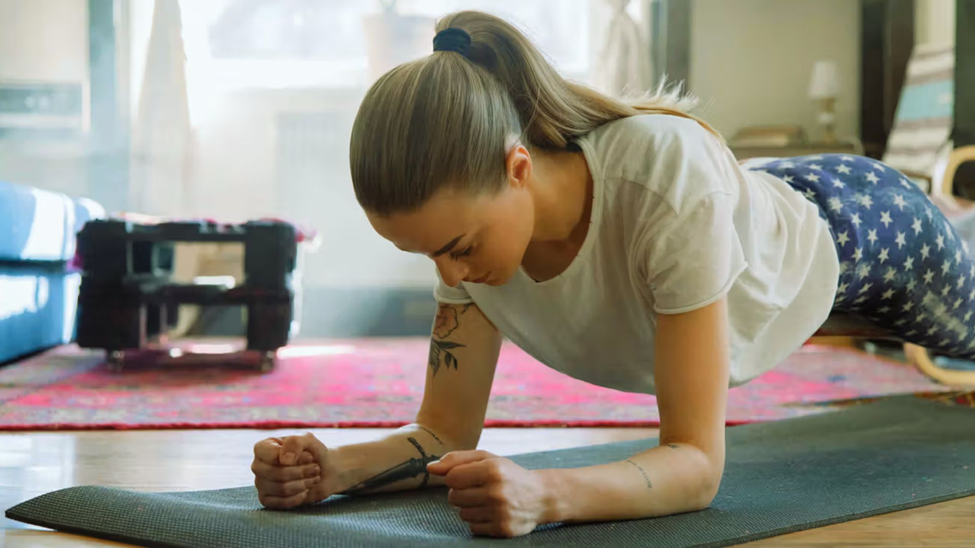 A woman in a white shirt and star-patterned leggings holds a plank pose on a yoga mat in a cozy living room. Natural light creates a calm atmosphere.