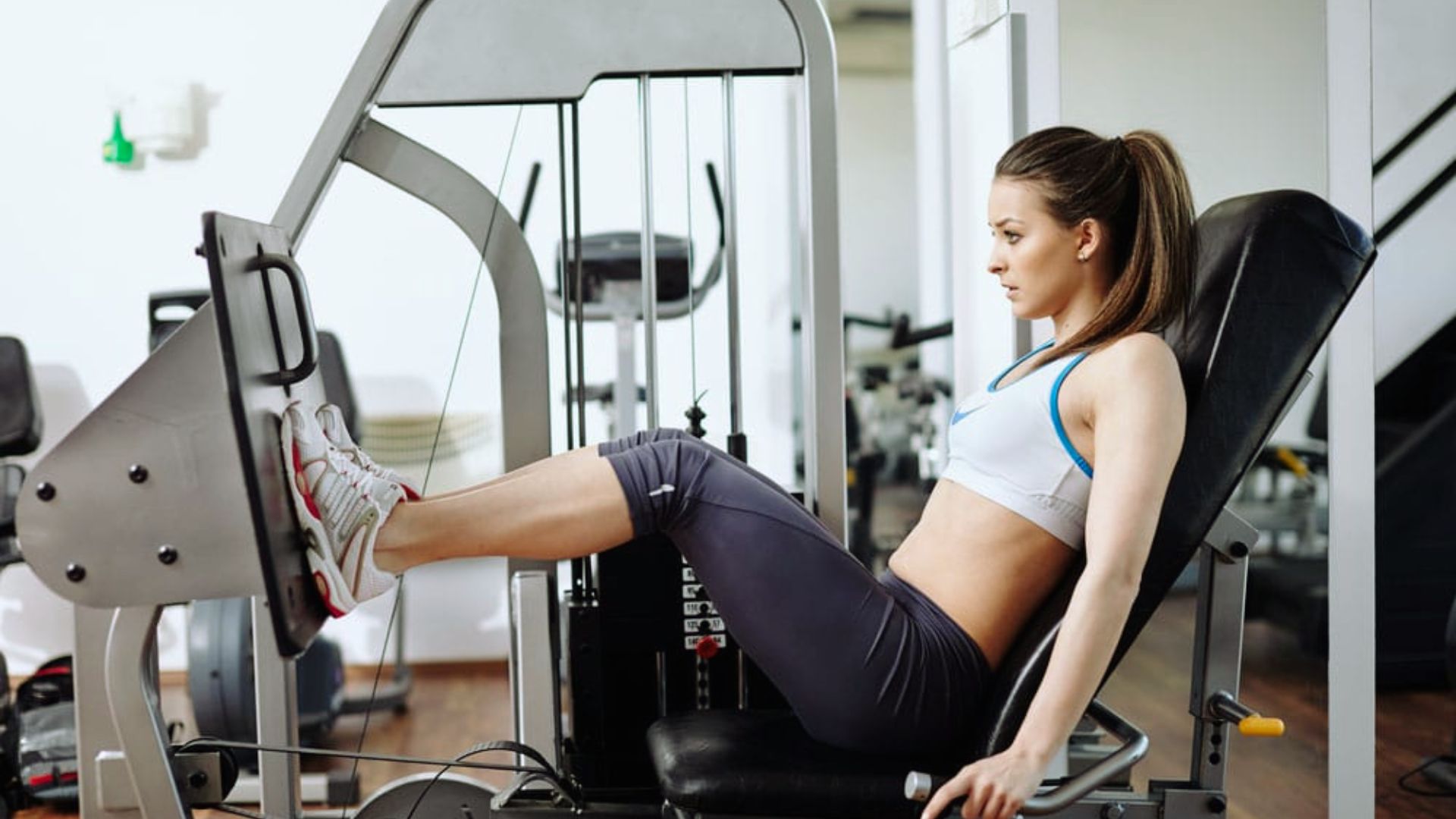 A woman in athletic wear uses a leg press machine in a gym, focused and determined. The setting is modern with exercise equipment visible.
