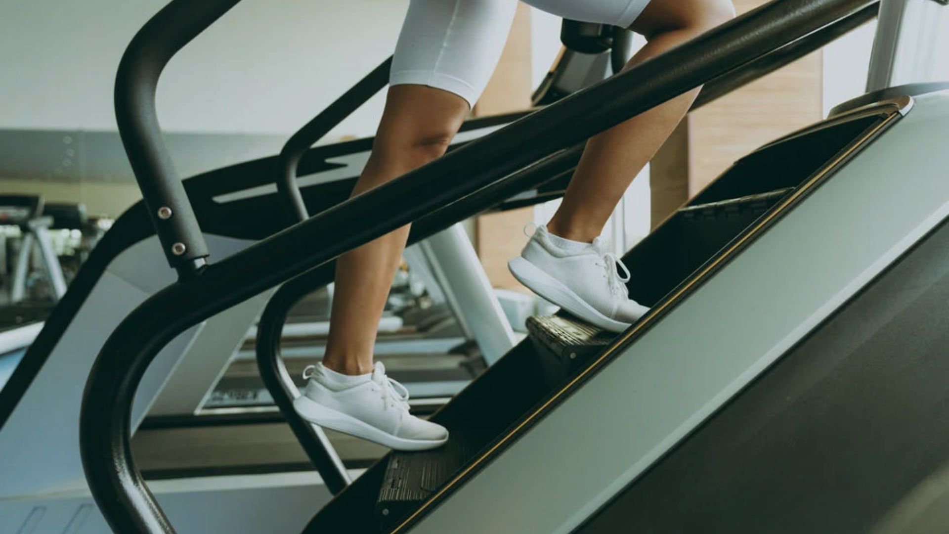 A person in white shorts and sneakers is using a stair climber at a modern gym. The focus is on the legs, emphasizing fitness and determination.