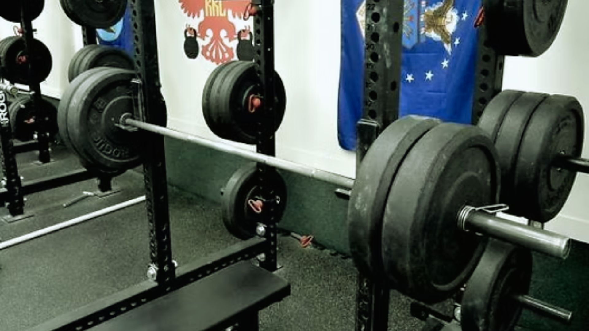 A weightlifting gym scene with a barbell loaded with heavy black plates on a squat rack. There are various weights stored on racks against a wall adorned with flags.